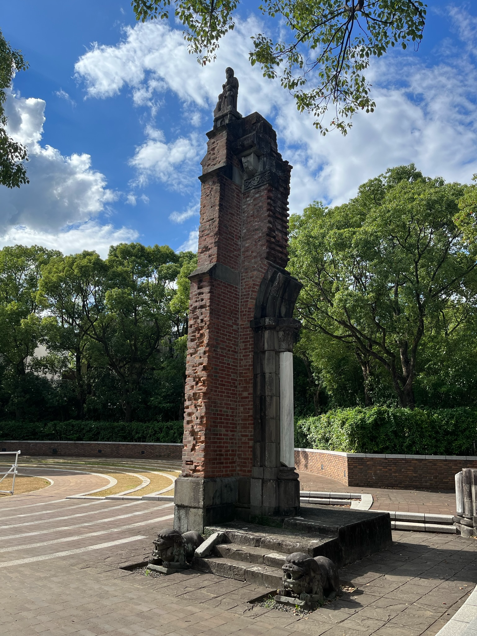 Ruins of a preserved brick bell tower from Urakami Cathedral in Nagasaki Peace Park, standing among trees and memorial grounds beneath a bright blue sky.