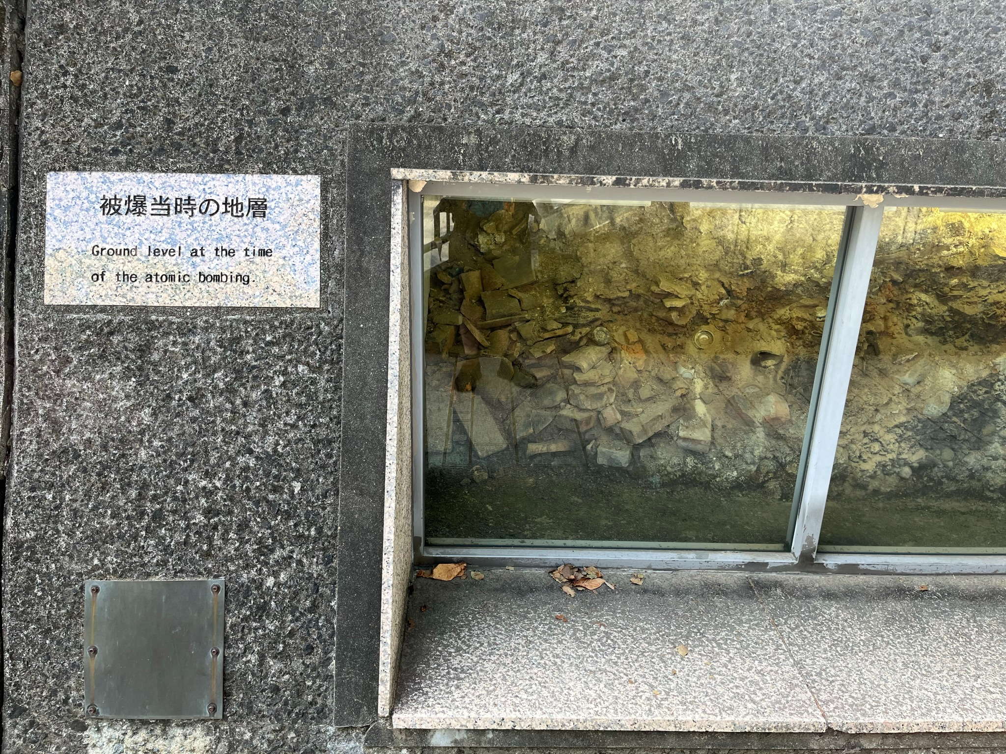 Preserved ground layer from the time of the Nagasaki atomic bombing, displayed behind glass with visible rubble, debris and a sign explaining the historic site.