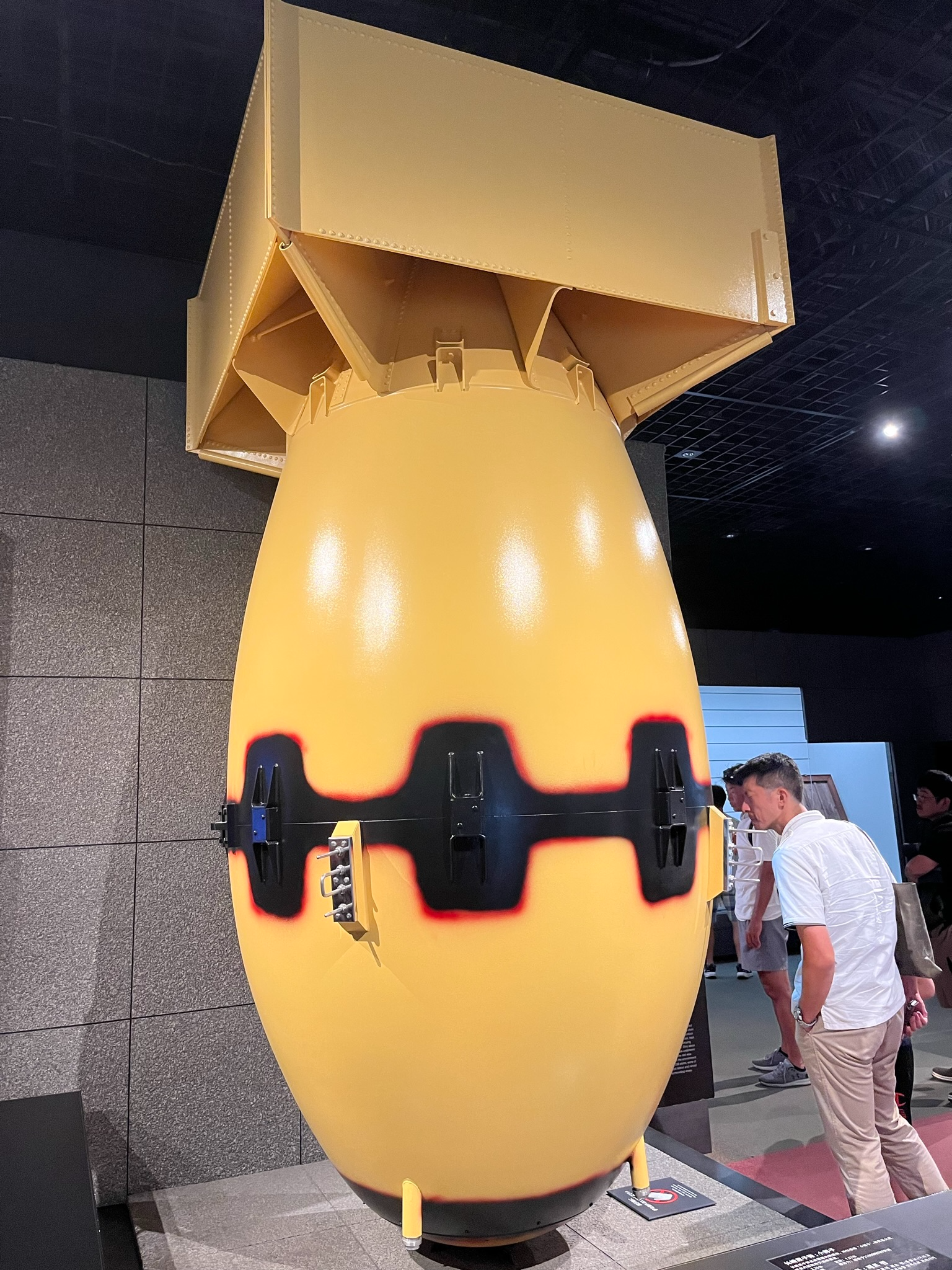 Full-scale replica of the "Fat Man" atomic bomb displayed at the Nagasaki Atomic Bomb Museum, with visitors standing beside the large yellow bomb model for scale.
