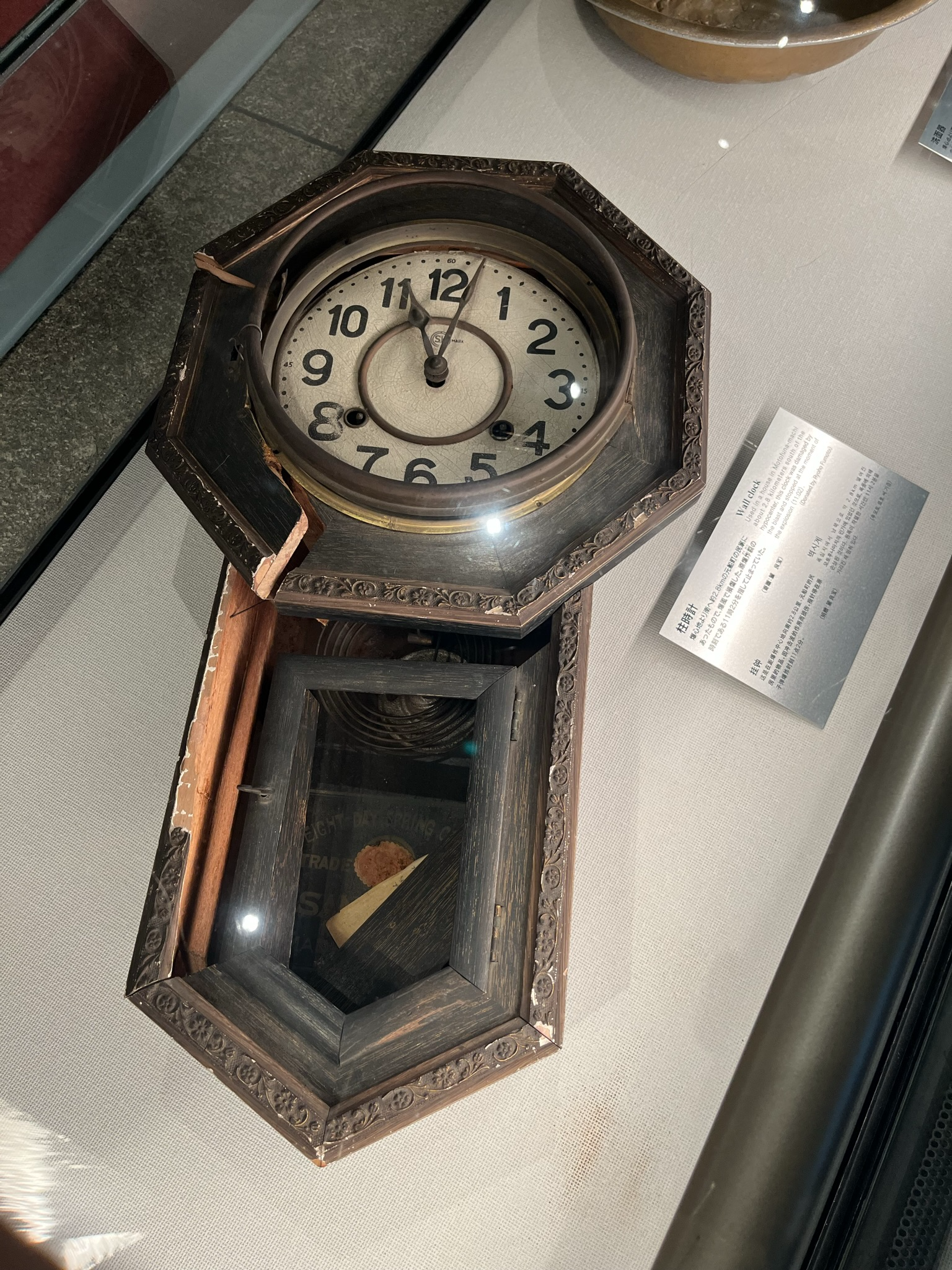 Damaged wall clock displayed at the Nagasaki Atomic Bomb Museum, with a cracked wooden case and stopped hands preserved as evidence of the 1945 atomic bombing.