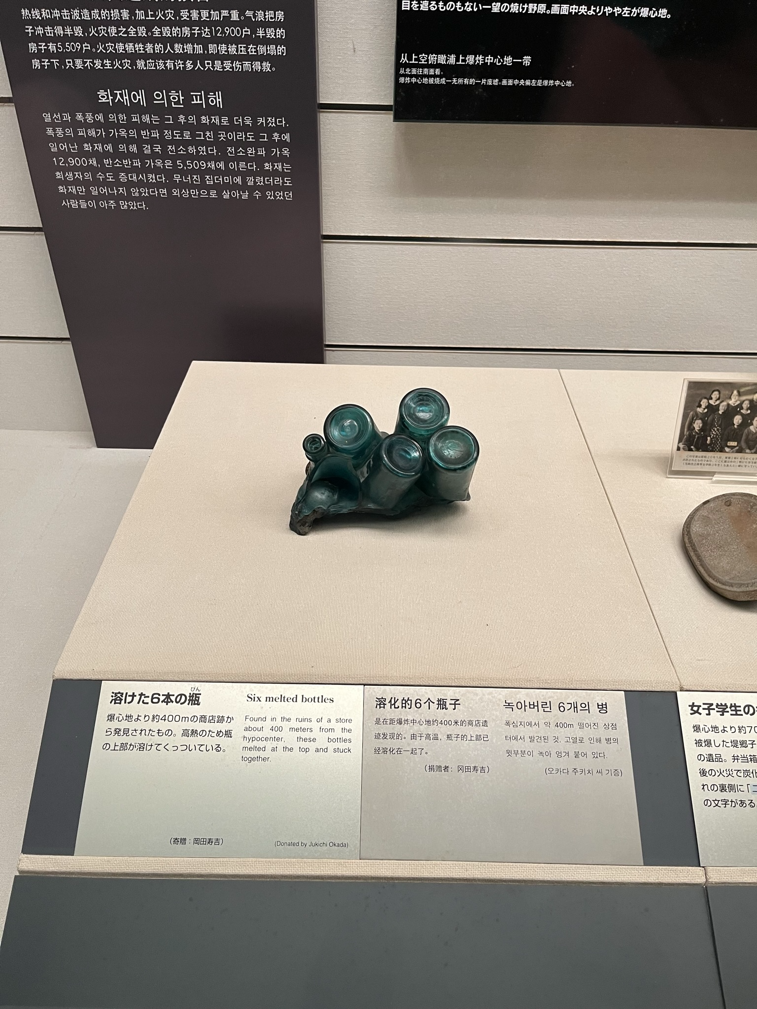 Six glass bottles melted and fused together by the heat of the Nagasaki atomic bombing, displayed in a museum case with explanatory information beside the artefact.