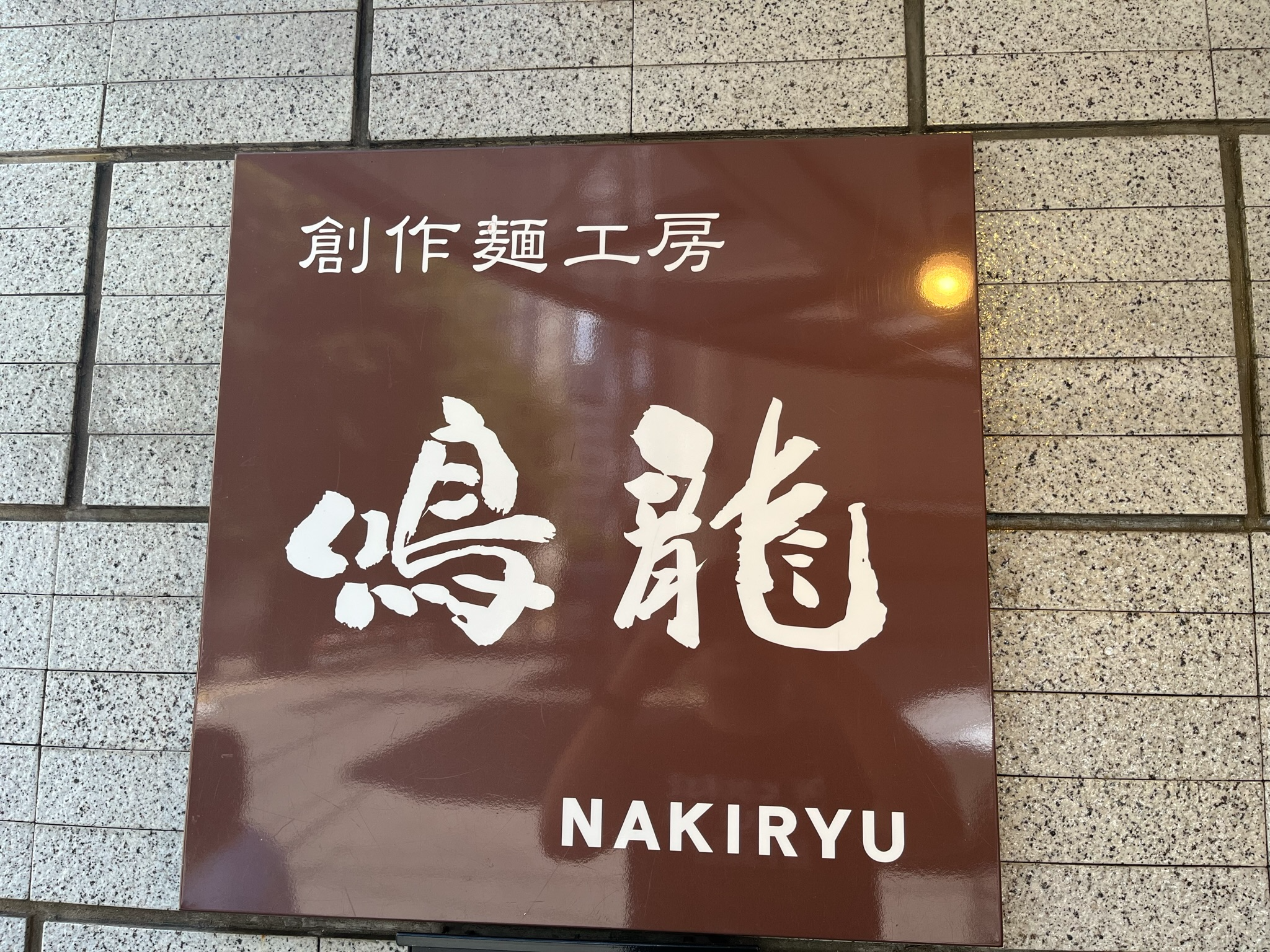 Sign for Nakiryu ramen restaurant in Tokyo, featuring Japanese calligraphy and the restaurant name in English on a brown panel mounted against a tiled wall.