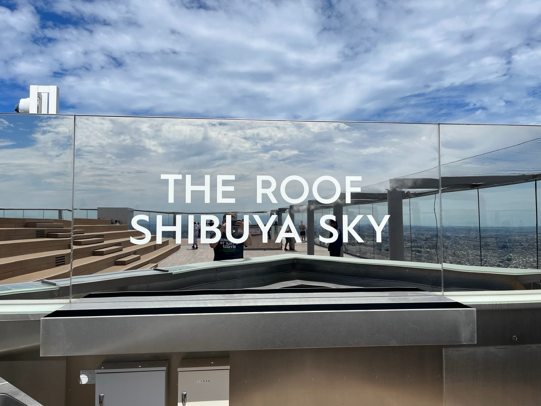 The Rooftop observation area at Shibuya Sky, featuring seating terraces, glass safety panels and sweeping views across Tokyo beneath dramatic cloud-filled skies.