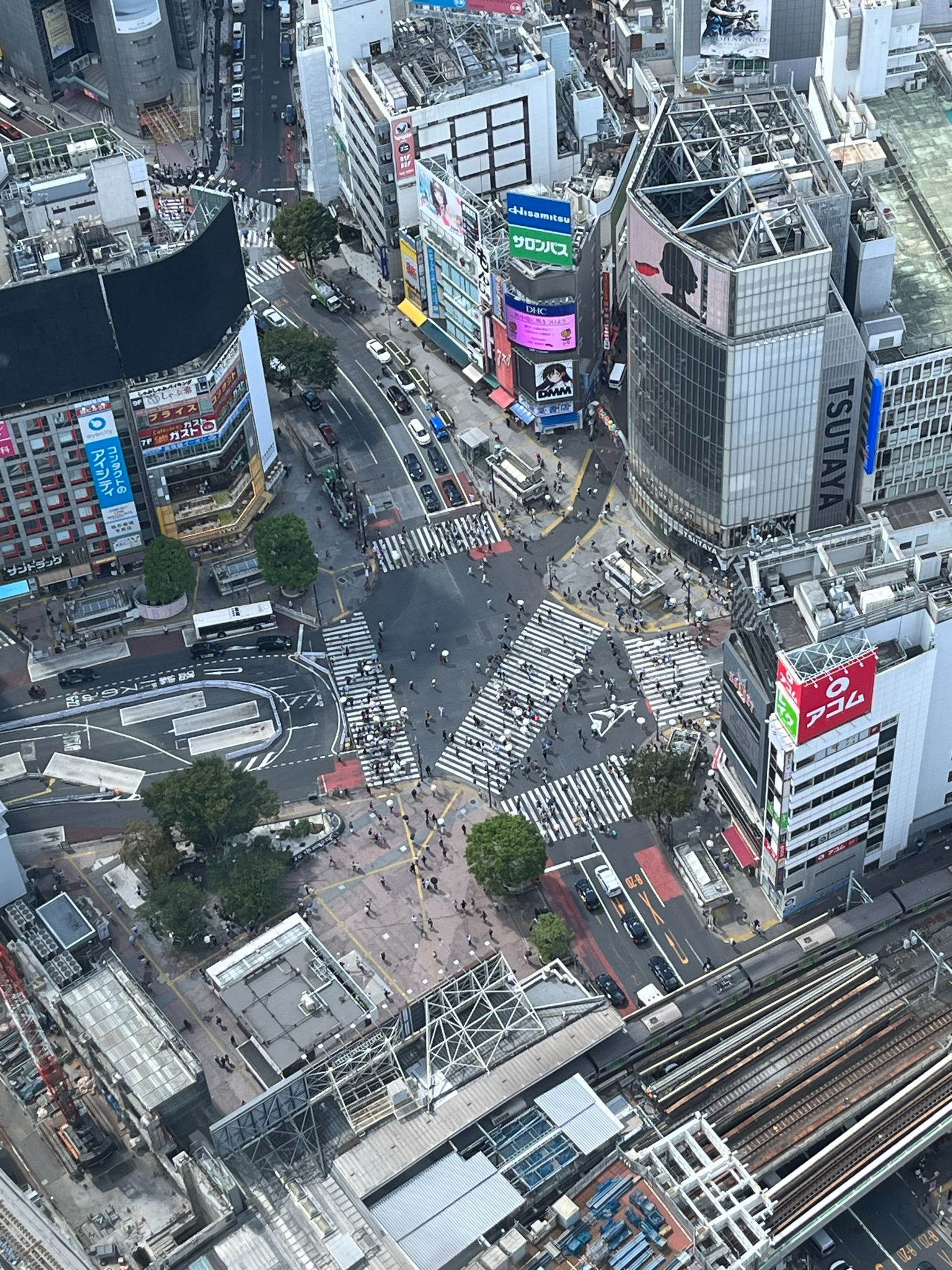 View from Shibuya Sky overlooking Shibuya Scramble Crossing, with pedestrians crossing from multiple directions surrounded by shops, offices, rail lines and city streets.