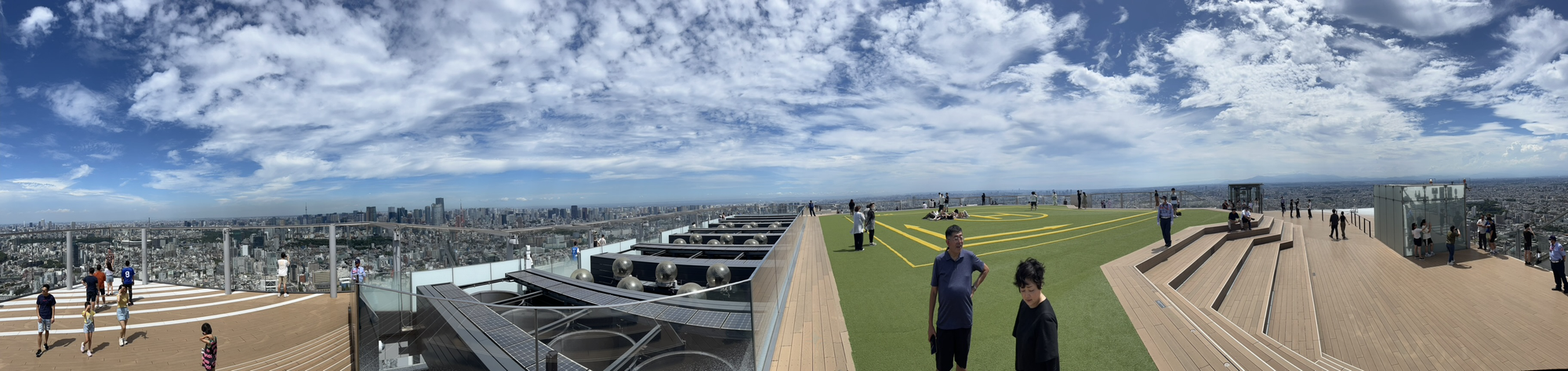 Panoramic view from Shibuya Sky's rooftop observation deck, with visitors enjoying open-air terraces and sweeping views across Tokyo beneath dramatic cloud-filled skies.