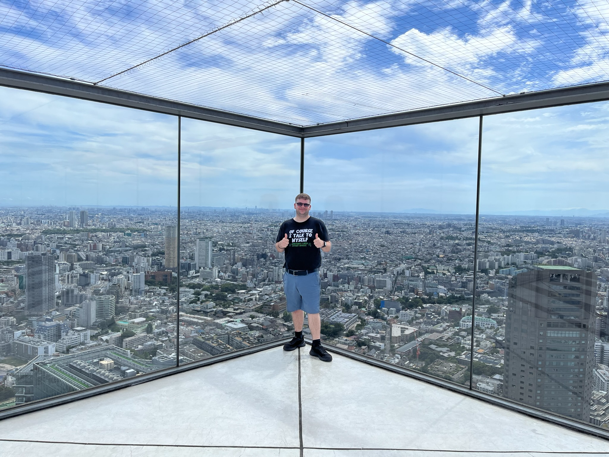 Visitor standing on the Shibuya Sky observation deck, surrounded by glass walls and sweeping panoramic views across Tokyo beneath a dramatic cloud-filled sky.