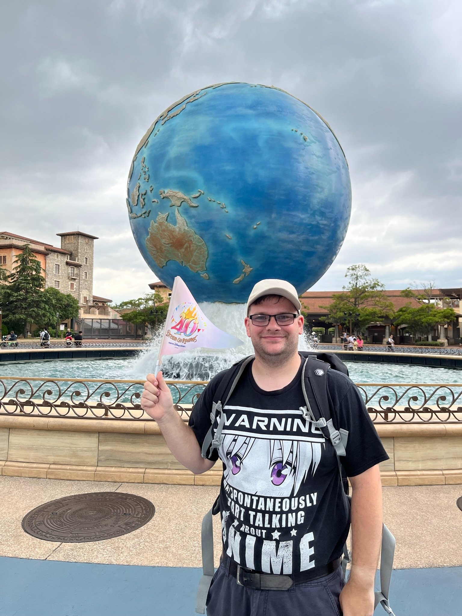 Visitor holding a Tokyo Disney Resort 40th Dream-Go-Round flag in front of the AquaSphere fountain at Tokyo DisneySea, with the iconic globe and park entrance behind.