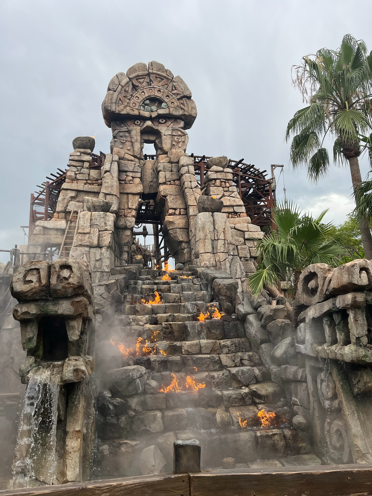 The entrance structure of Raging Spirits at Tokyo DisneySea, featuring ancient stone ruins, carved faces, flames, waterfalls and tropical vegetation beneath dark stormy skies.