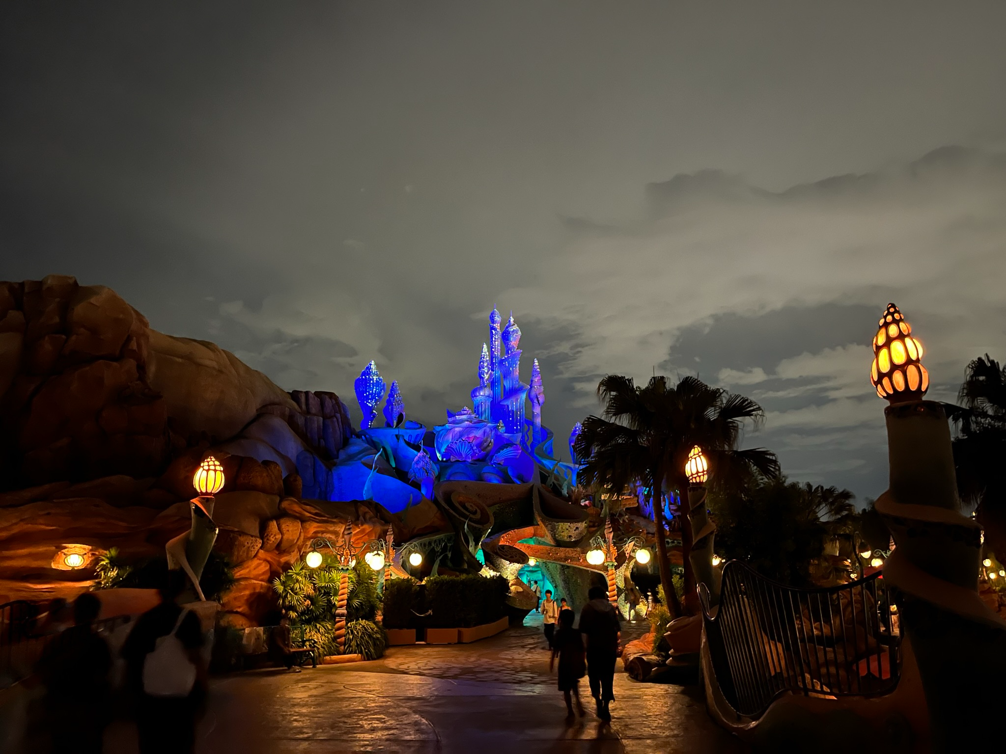 Mermaid Lagoon at Tokyo DisneySea illuminated at night, featuring King Triton's colourful palace, glowing lanterns, rocky landscapes and visitors walking through the fantasy-themed area.