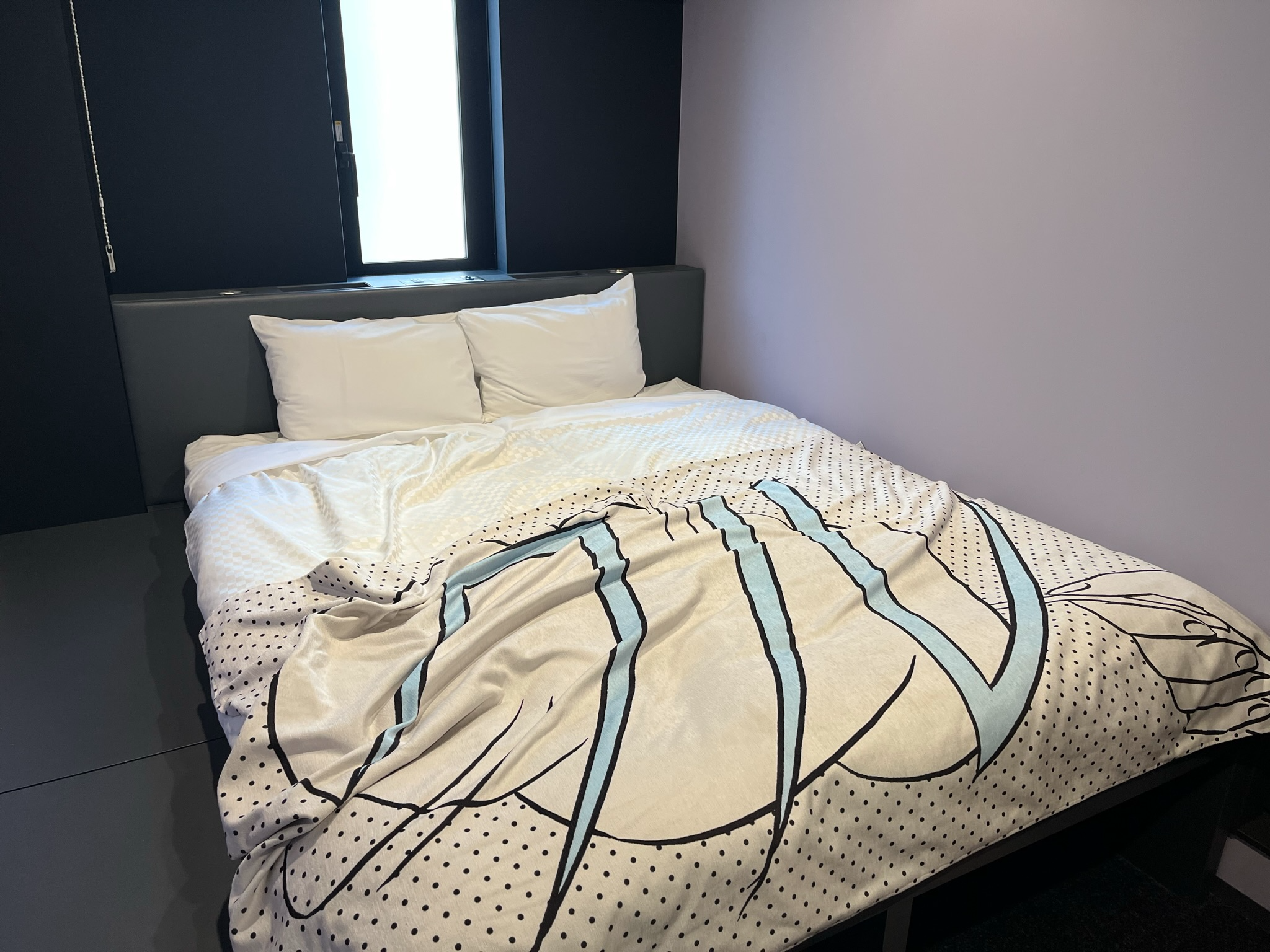 Compact hotel room with a double bed, black furnishings and anime-themed bedding, illuminated by natural light from a small window above the headboard.