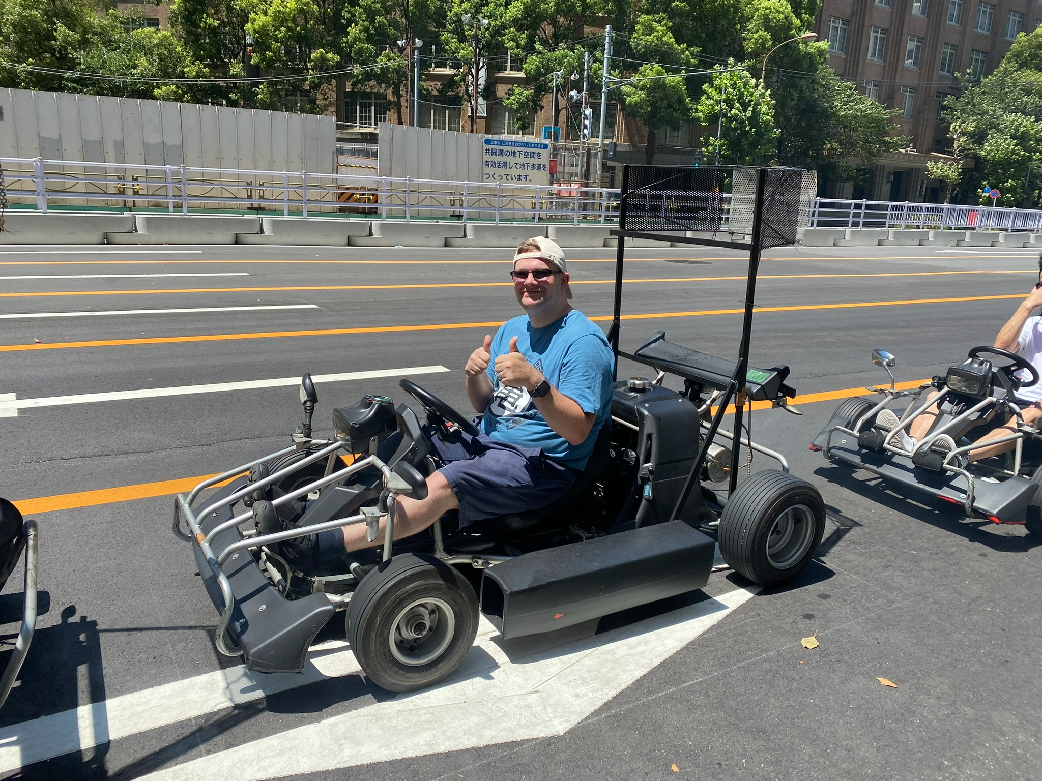 Participant giving a thumbs up while seated in a street go-kart on a Tokyo roadway, with other go-karts, city streets and buildings visible on a sunny day.