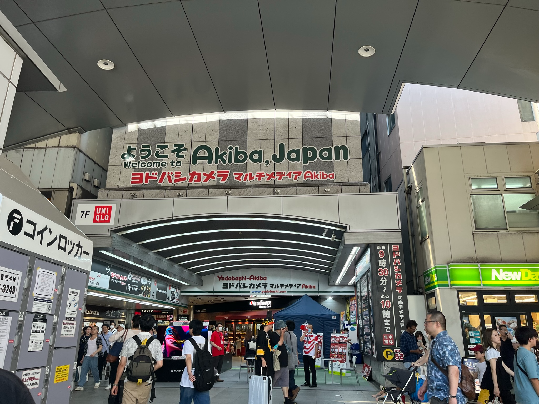 Entrance to Yodobashi Camera Multimedia Akiba in Akihabara, with shoppers passing beneath a large "Welcome to Akiba, Japan" sign at one of Tokyo's busiest retail destinations.
