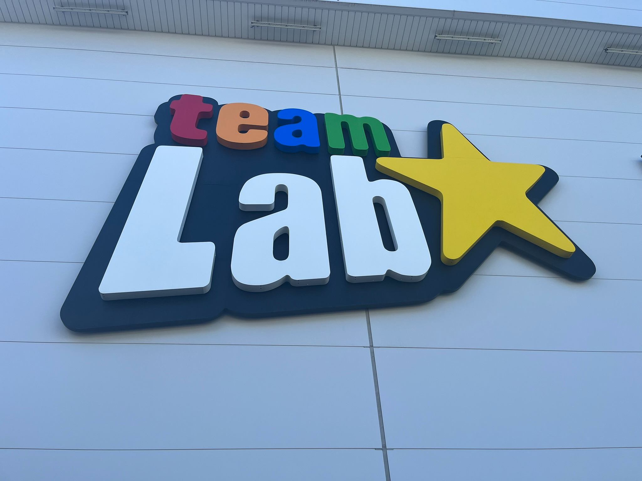 Large TeamLab sign mounted on the exterior of TeamLab Planets Tokyo, featuring colourful lettering and a yellow star against a modern white building facade.