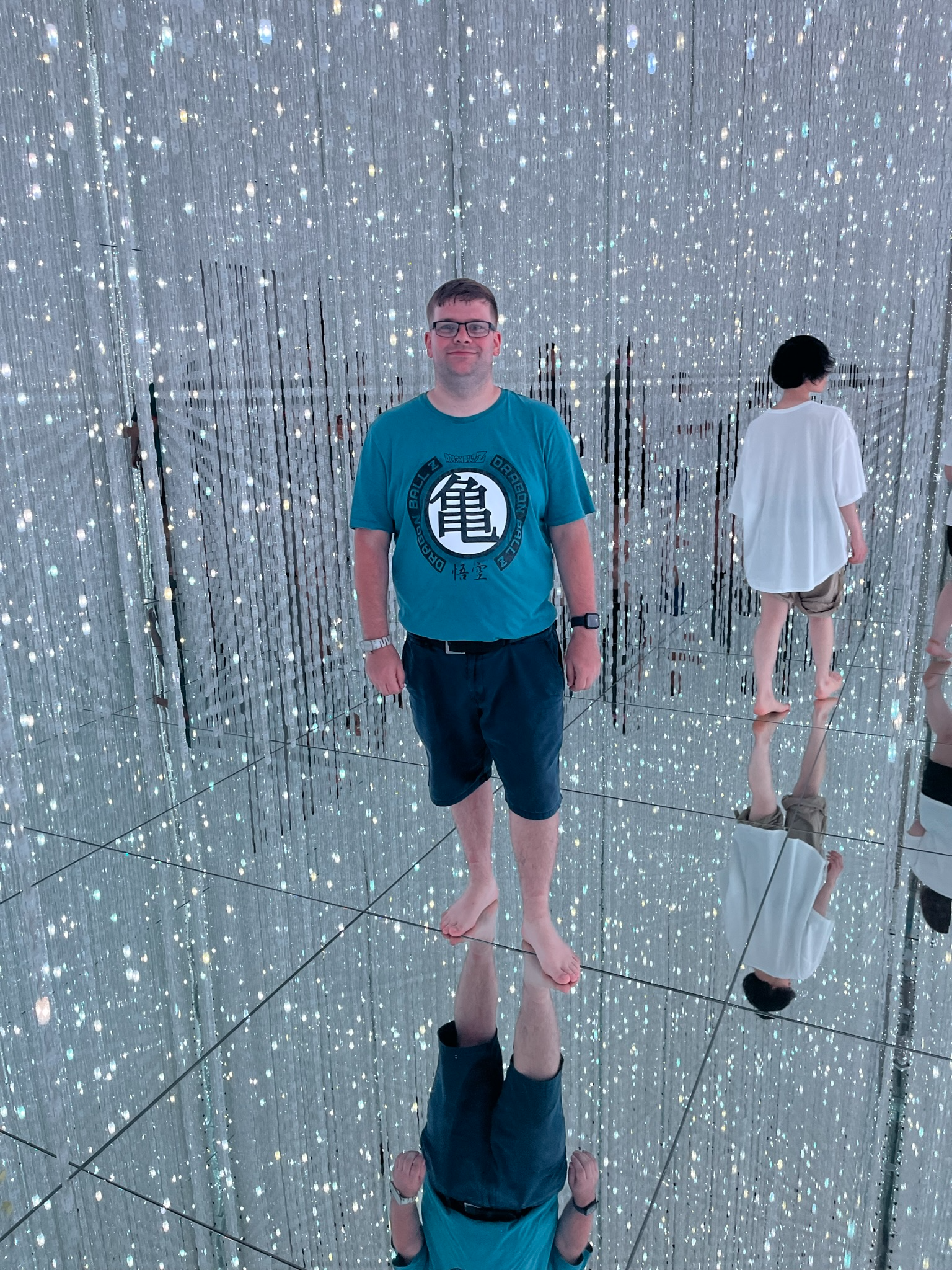 Visitor standing barefoot inside TeamLab Planets' mirrored light installation, surrounded by thousands of sparkling suspended lights reflected in every direction.