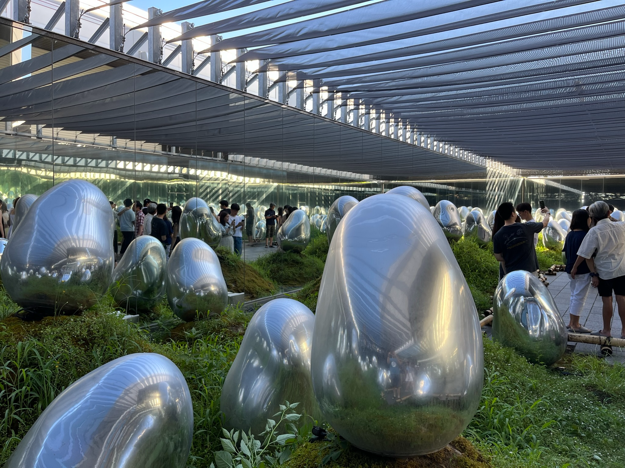 Reflective silver egg-shaped sculptures among moss and grass in TeamLab Planets Tokyo's outdoor garden installation, with mirrored walls and visitors exploring the artwork.
