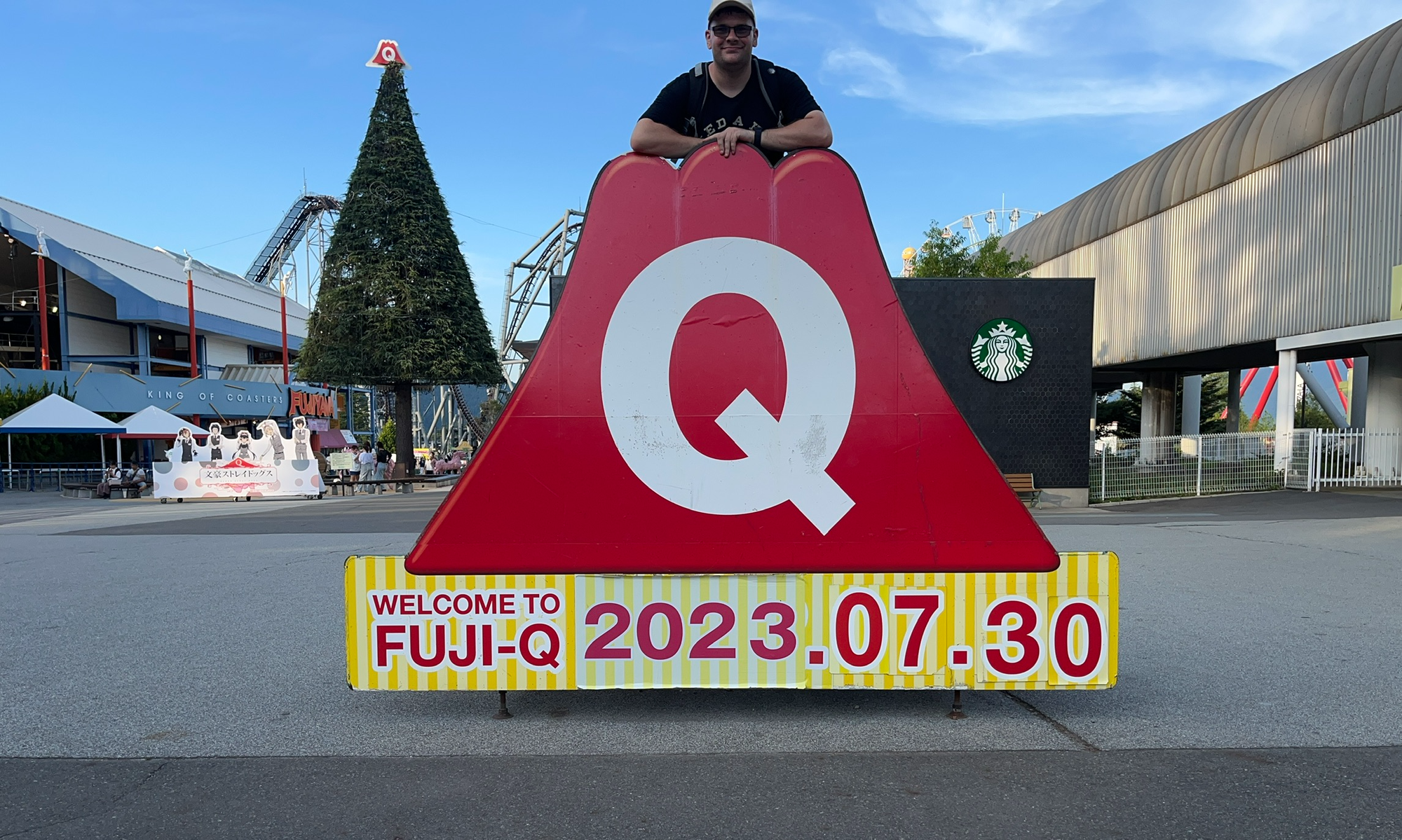 Visitor posing behind a large Fuji-Q Highland sign, with roller coaster tracks, park attractions and a Starbucks store visible in the background under clear evening skies.