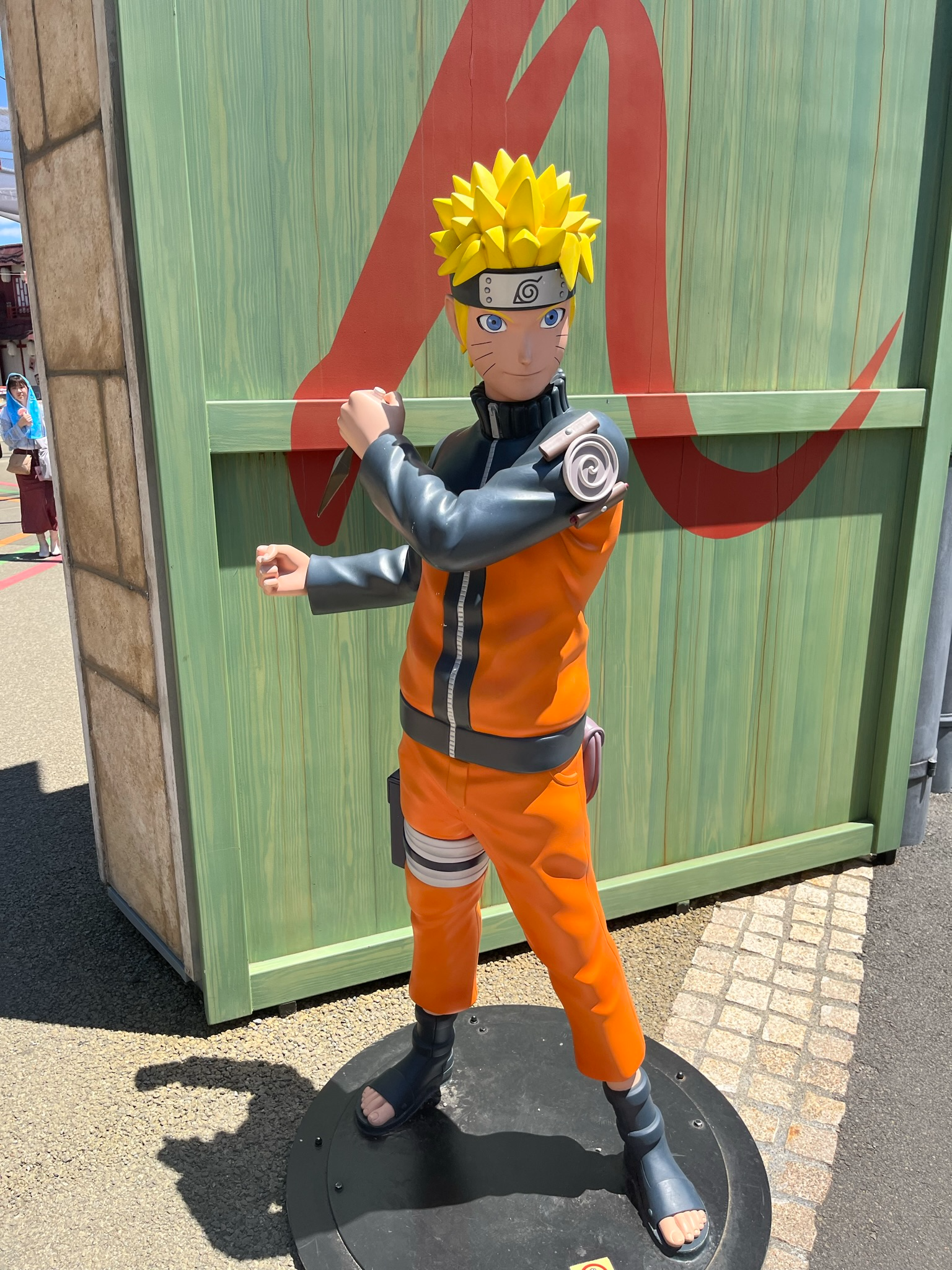 Statue of Naruto Uzumaki from the Naruto series at Fuji-Q Highland, showing the character in an action pose against a themed backdrop featuring the Hidden Leaf Village symbol.
