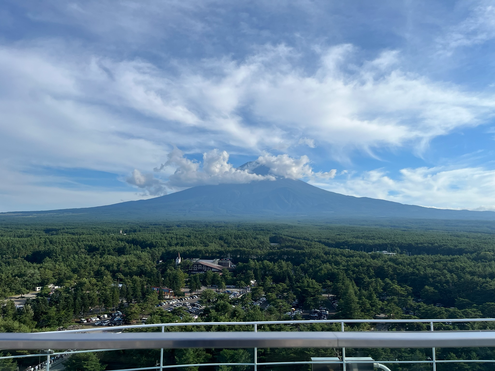 Mount Fuji rises above a vast forest landscape viewed from Fujiyama Tower, with dramatic white clouds drifting around the summit beneath a bright blue summer sky.