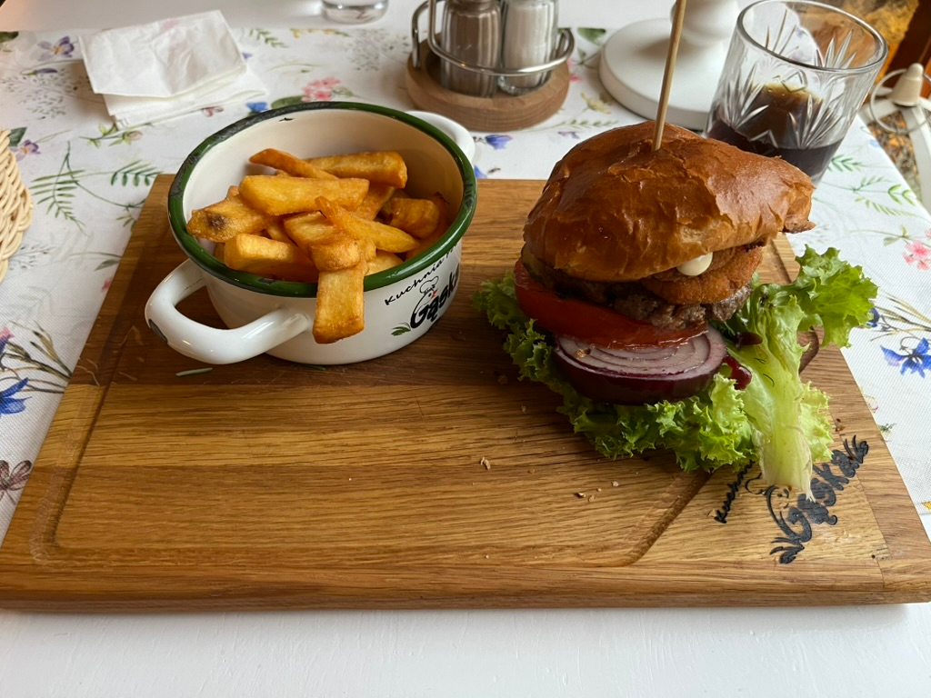Burger with lettuce, tomato, red onion, and a fried topping, served with thick-cut fries on a wooden board.