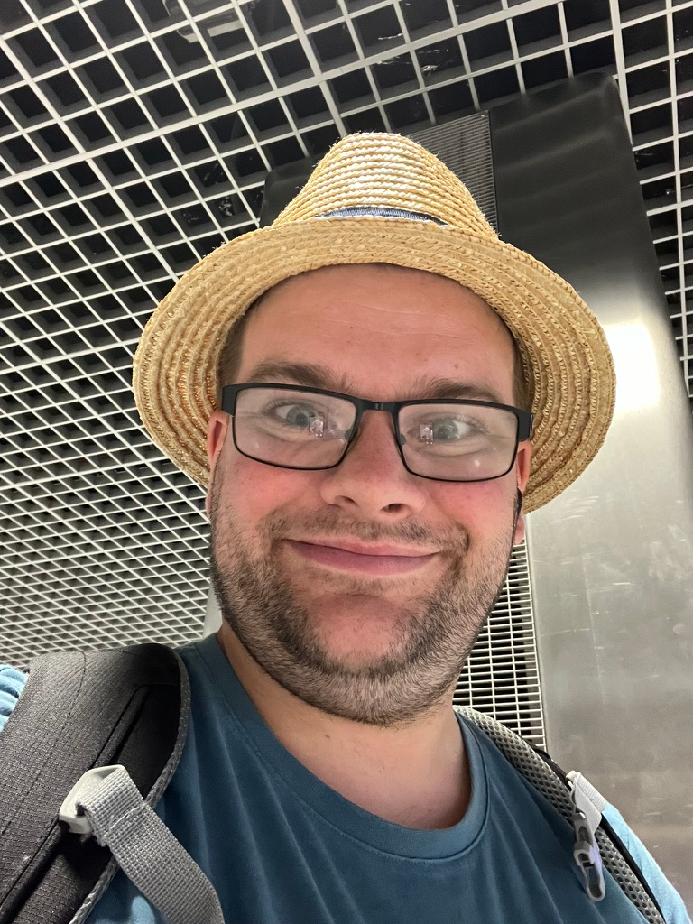 Close-up selfie of a person wearing glasses and a straw hat, with backpack straps visible indoors beneath a grid-pattern ceiling.