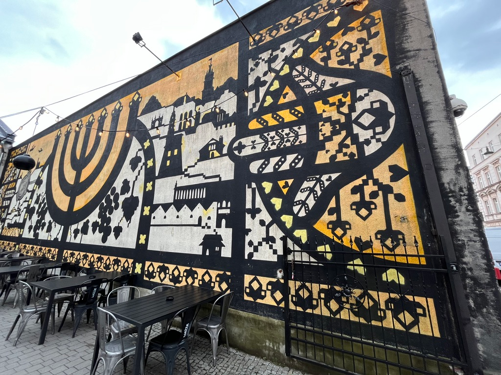 Large black, white, and gold mural at the Galicia Jewish Museum in Kraków, featuring a menorah, city skyline silhouettes, and intricate geometric patterns above an outdoor seating area.