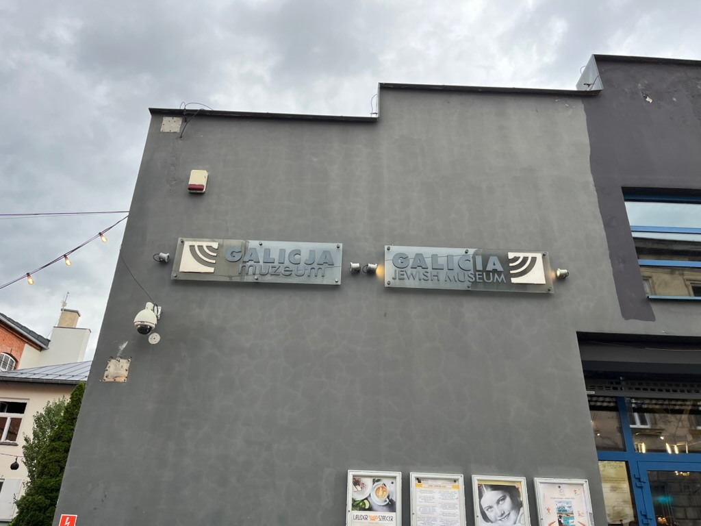 Exterior wall of the Galicia Jewish Museum in Kraków, featuring bilingual museum signs on a grey building beneath an overcast sky.