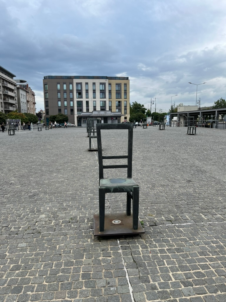 Metal chair sculptures arranged across a large cobbled square in Kraków, with modern buildings and a cloudy sky in the background.