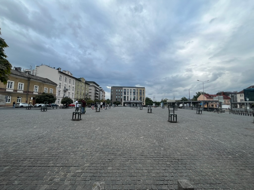 Wide view of Ghetto Heroes Square in Kraków, with rows of metal chair memorials spread across a large cobbled plaza beneath a dramatic cloudy sky.