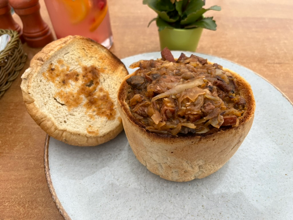 Open bread bowl filled with traditional Polish bigos, a hearty stew of sauerkraut, cabbage and mixed meats, served on a ceramic plate with the bread lid alongside.