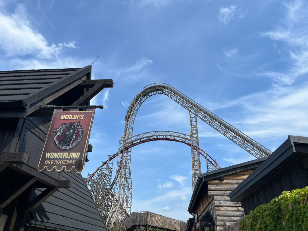 View of the Zadra roller coaster at Energylandia, with its towering wooden support structure rising above themed buildings beneath a bright blue sky.