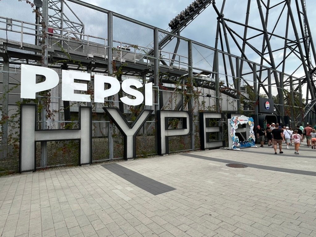Large "Pepsi Hyperion" sign beside the entrance to Hyperion roller coaster at Energylandia, with the towering black steel track and trains visible overhead.