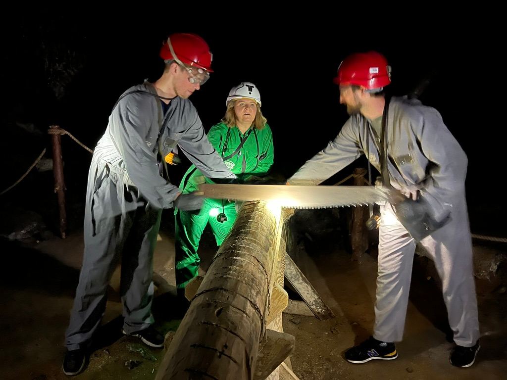 Visitors wearing mining overalls, helmets and headlamps use a large two-person saw to cut a log during a hands-on activity inside the Wieliczka Salt Mine.