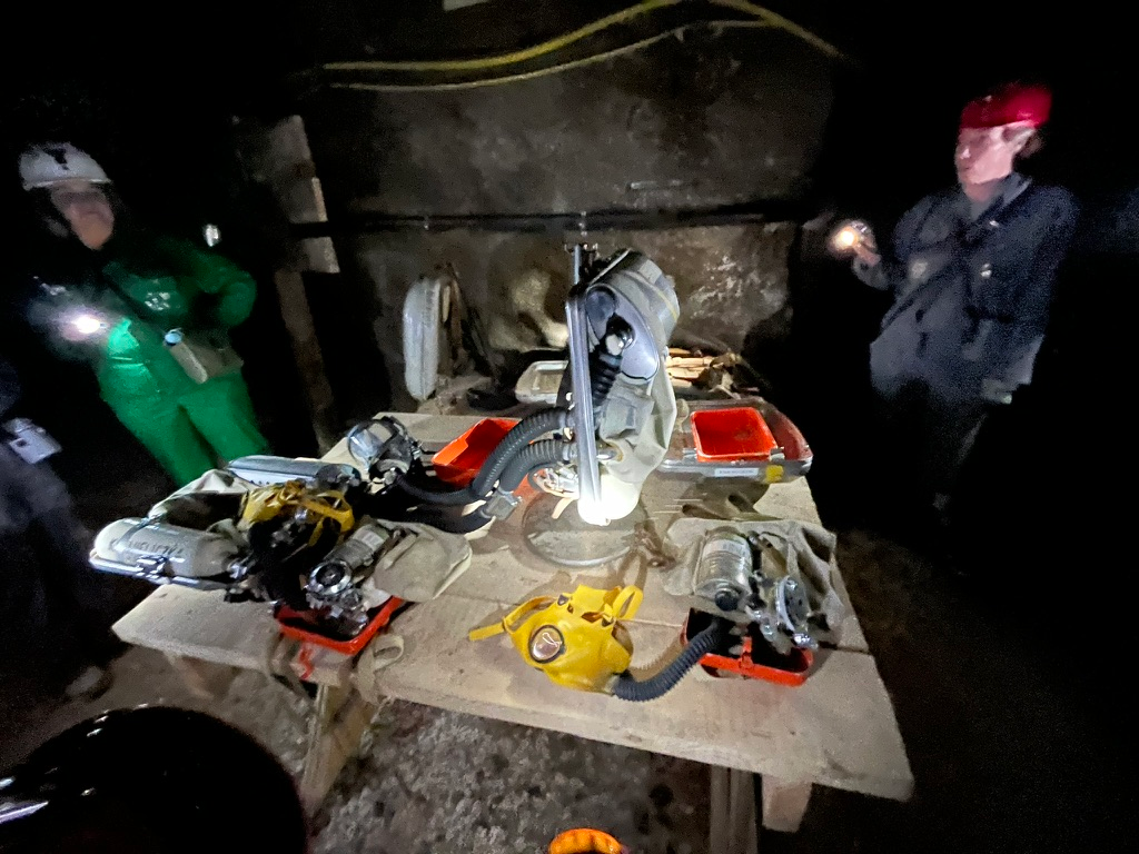 Table displaying historic mining and rescue equipment inside the Wieliczka Salt Mine, including breathing apparatus, helmets, gas masks and lamps, illuminated by visitors' headlamps.