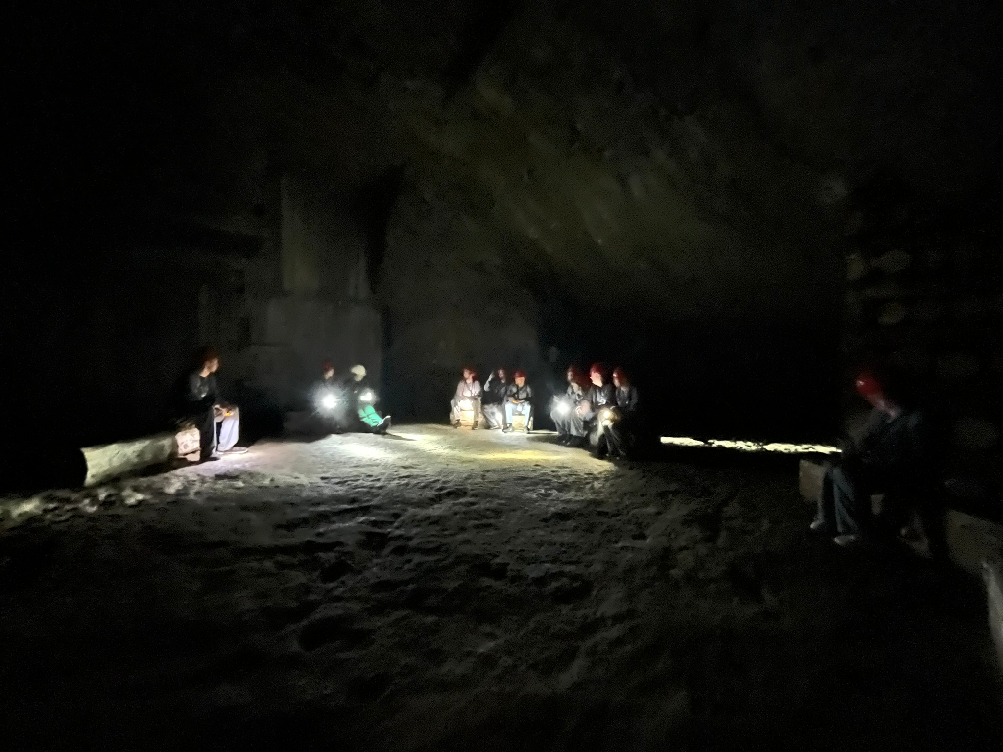 Visitors wearing helmets and headlamps sit around a large underground chamber inside the Wieliczka Salt Mine, illuminated only by pools of light in the darkness.