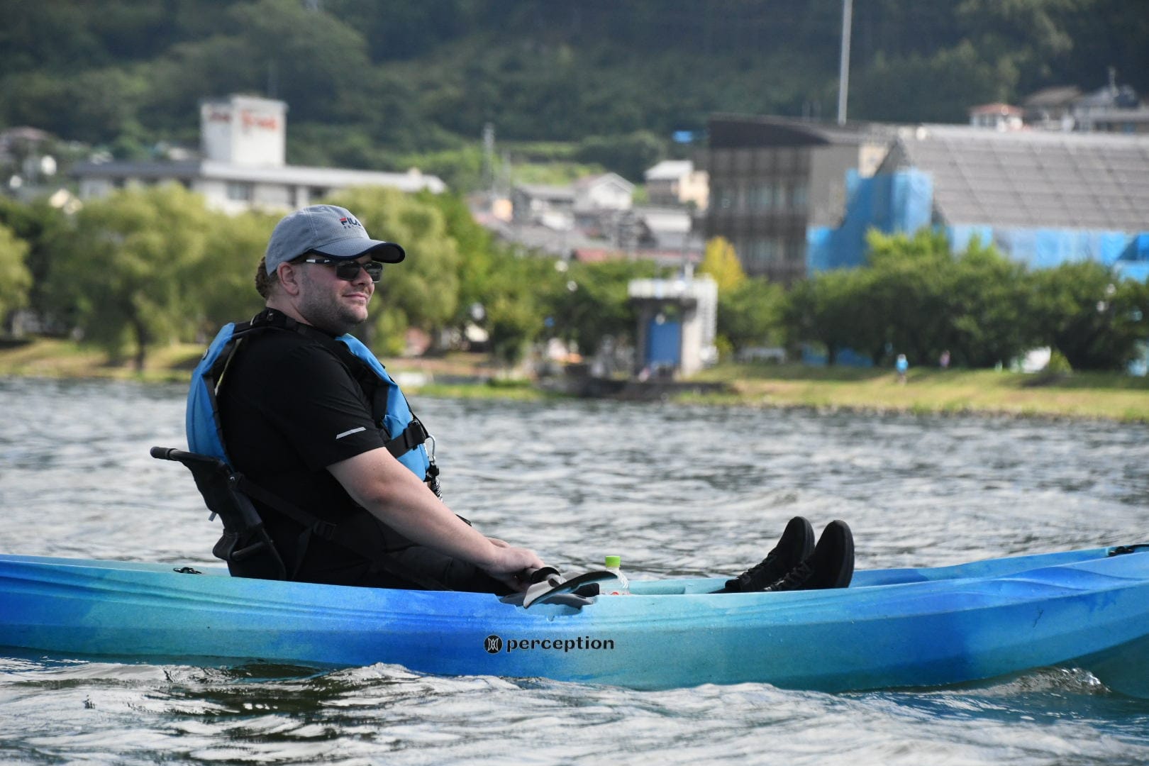 Person kayaking on Lake Suwa in a blue sit-on-top kayak, wearing a life jacket and cap, with the lakeshore, trees and buildings visible in the background.