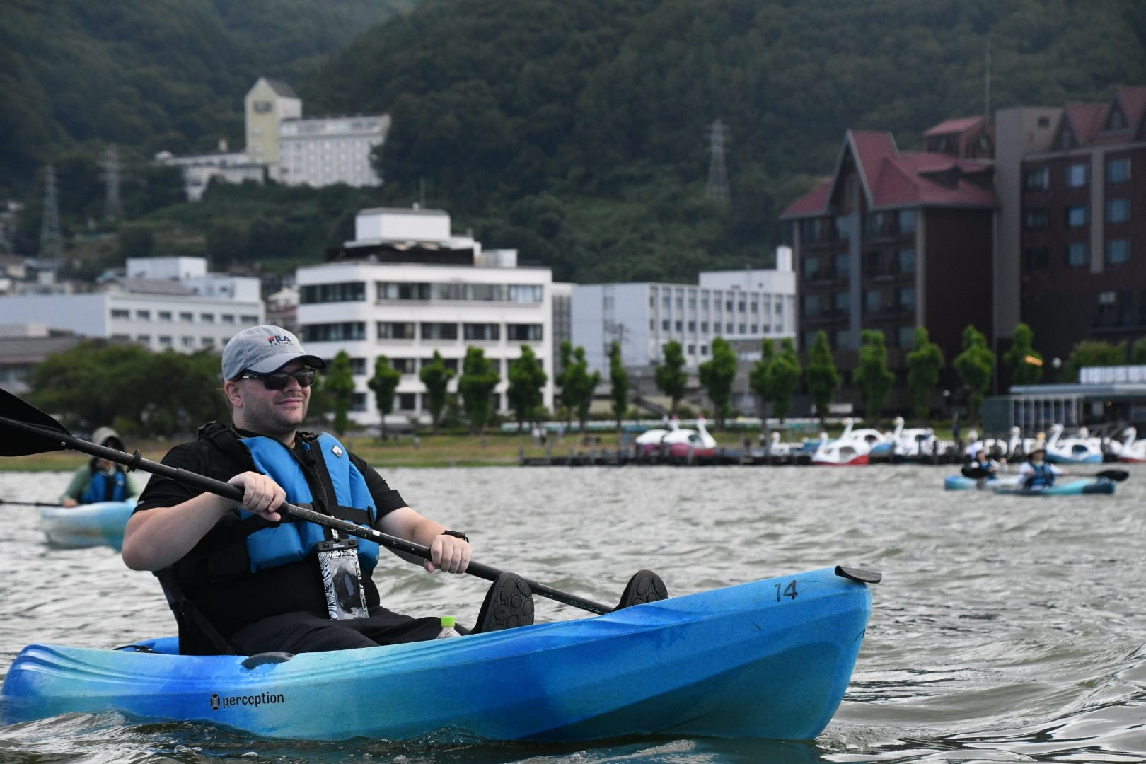 Person paddling a blue kayak on Lake Suwa, wearing a life jacket and cap, with lakeside hotels, swan boats and forested hills visible along the shoreline.