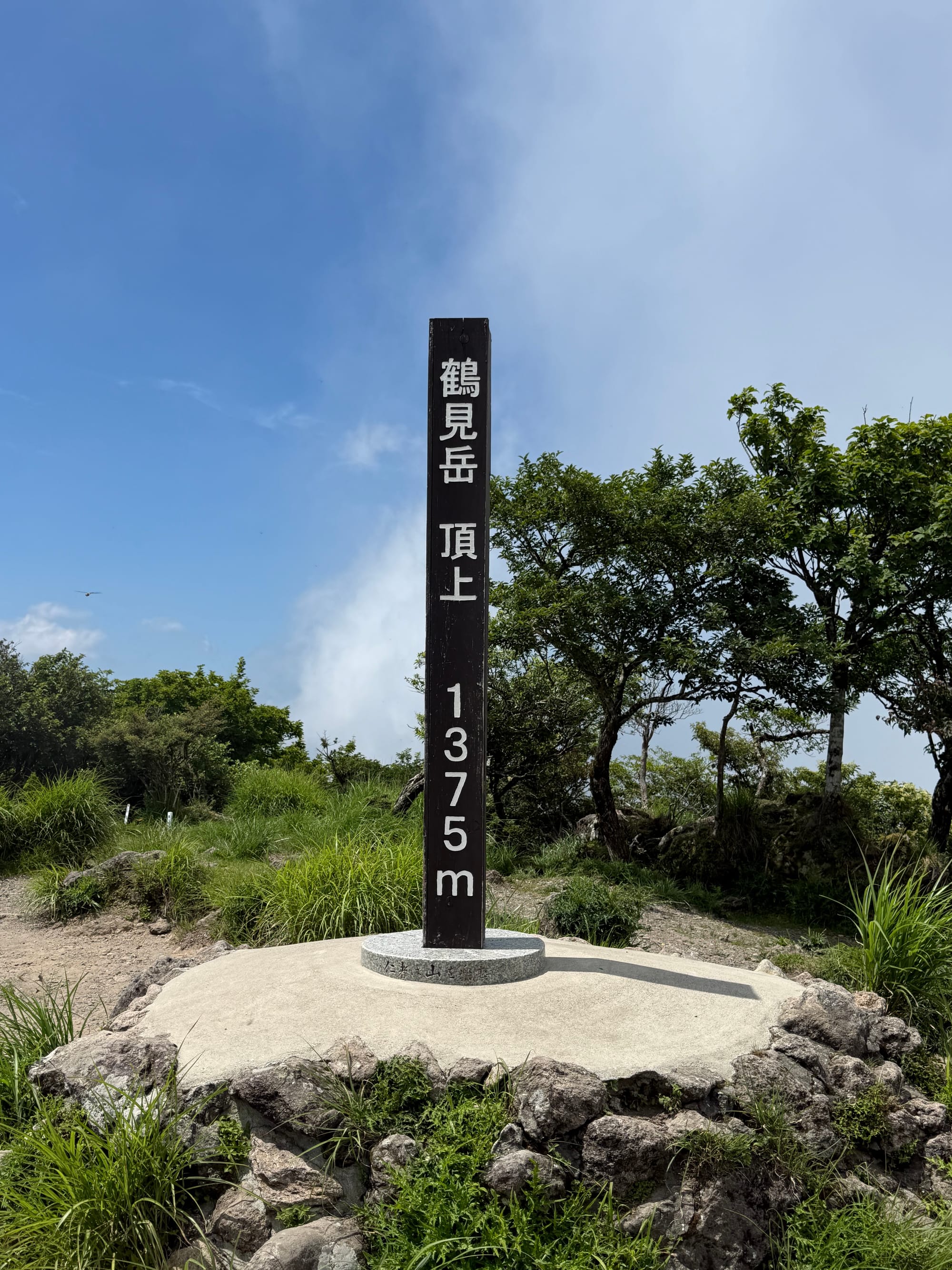 Summit marker on Mount Tsurumi showing an elevation of 1,375 metres, surrounded by grass, rocks and mountain vegetation beneath a bright summer sky.