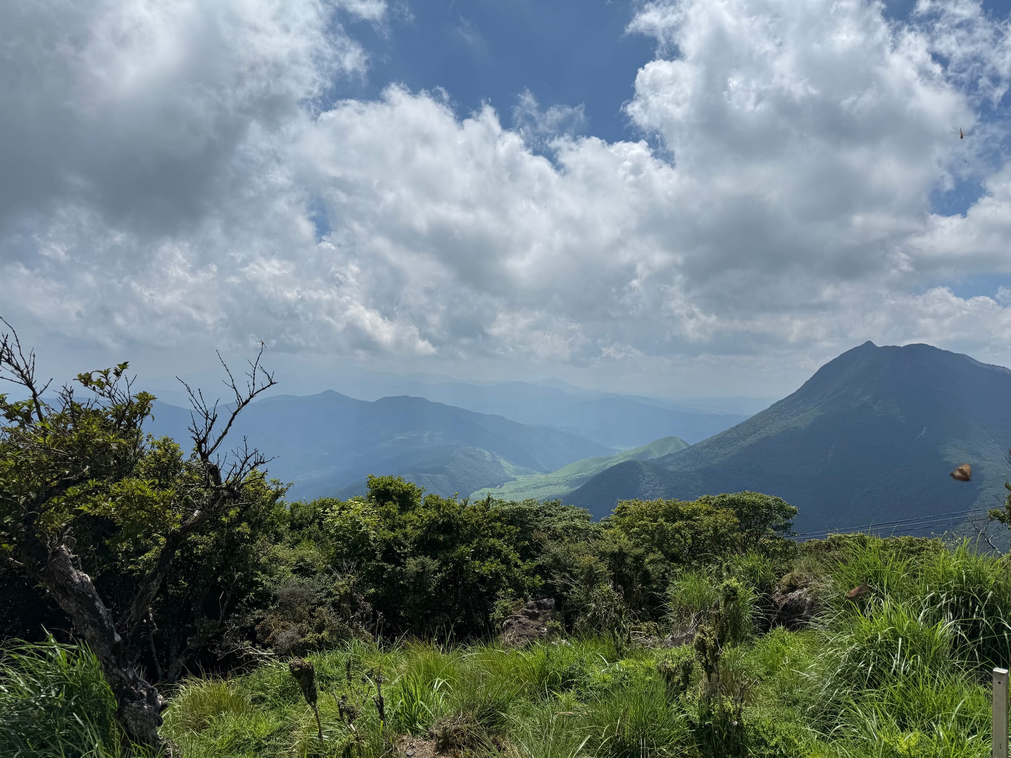 Mountain landscape viewed from Mount Tsurumi, featuring forested slopes, rolling peaks and valleys beneath dramatic clouds and patches of sunlight.