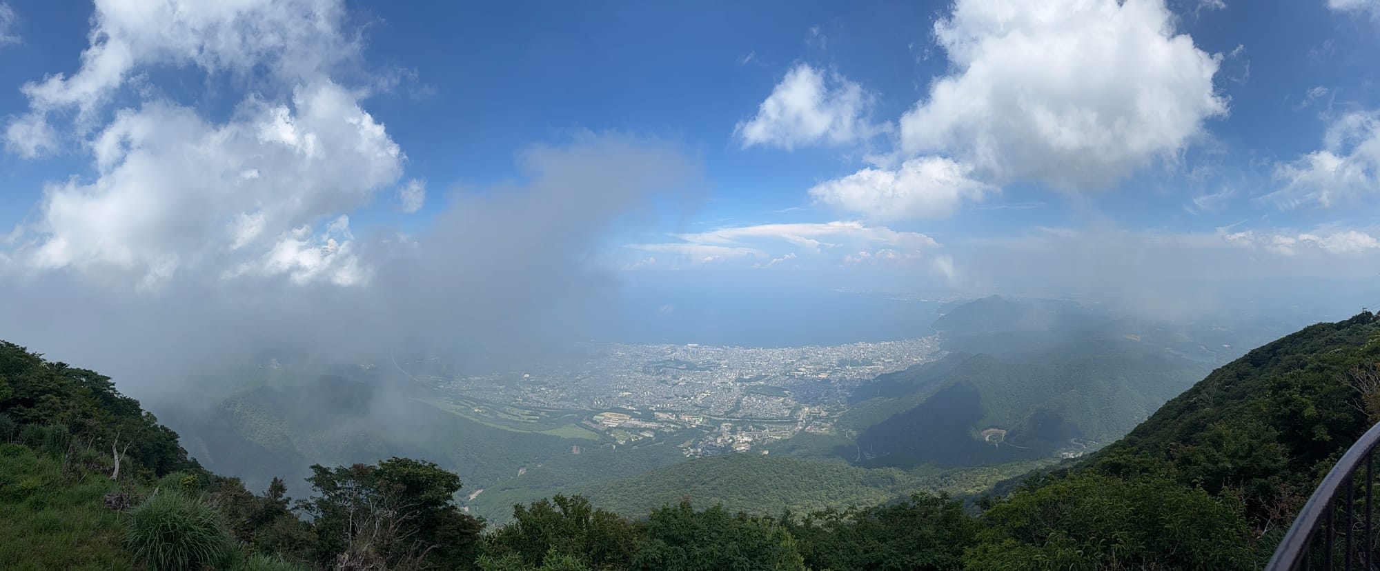 Panoramic view from Mount Tsurumi overlooking Beppu and Beppu Bay, with forested slopes, drifting clouds and patches of sunlight breaking through above the city.