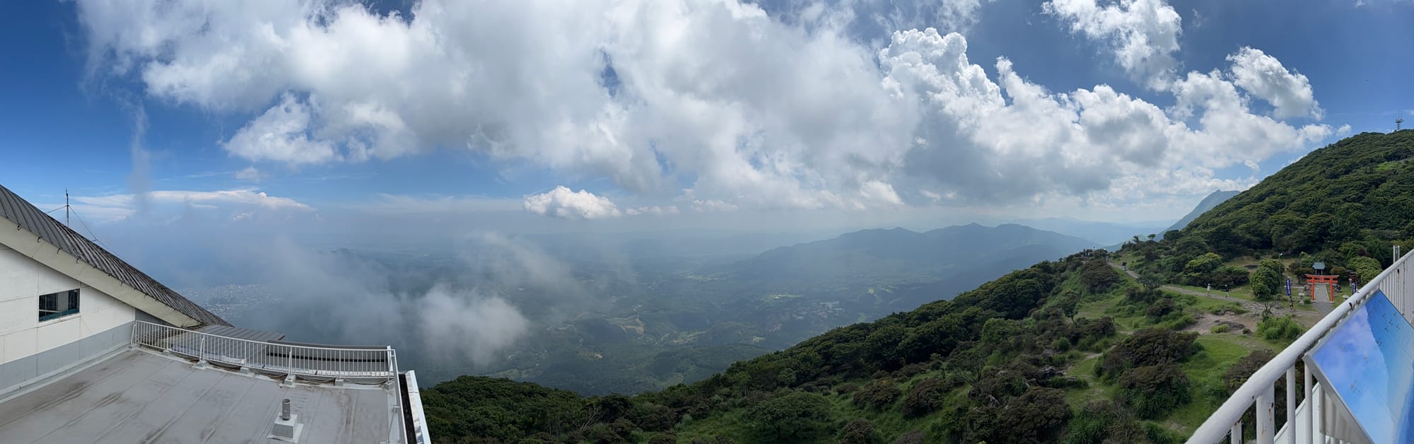 Panoramic view from Mount Tsurumi's ropeway summit station, overlooking forested valleys, mountain ridges and drifting clouds beneath a bright blue summer sky.