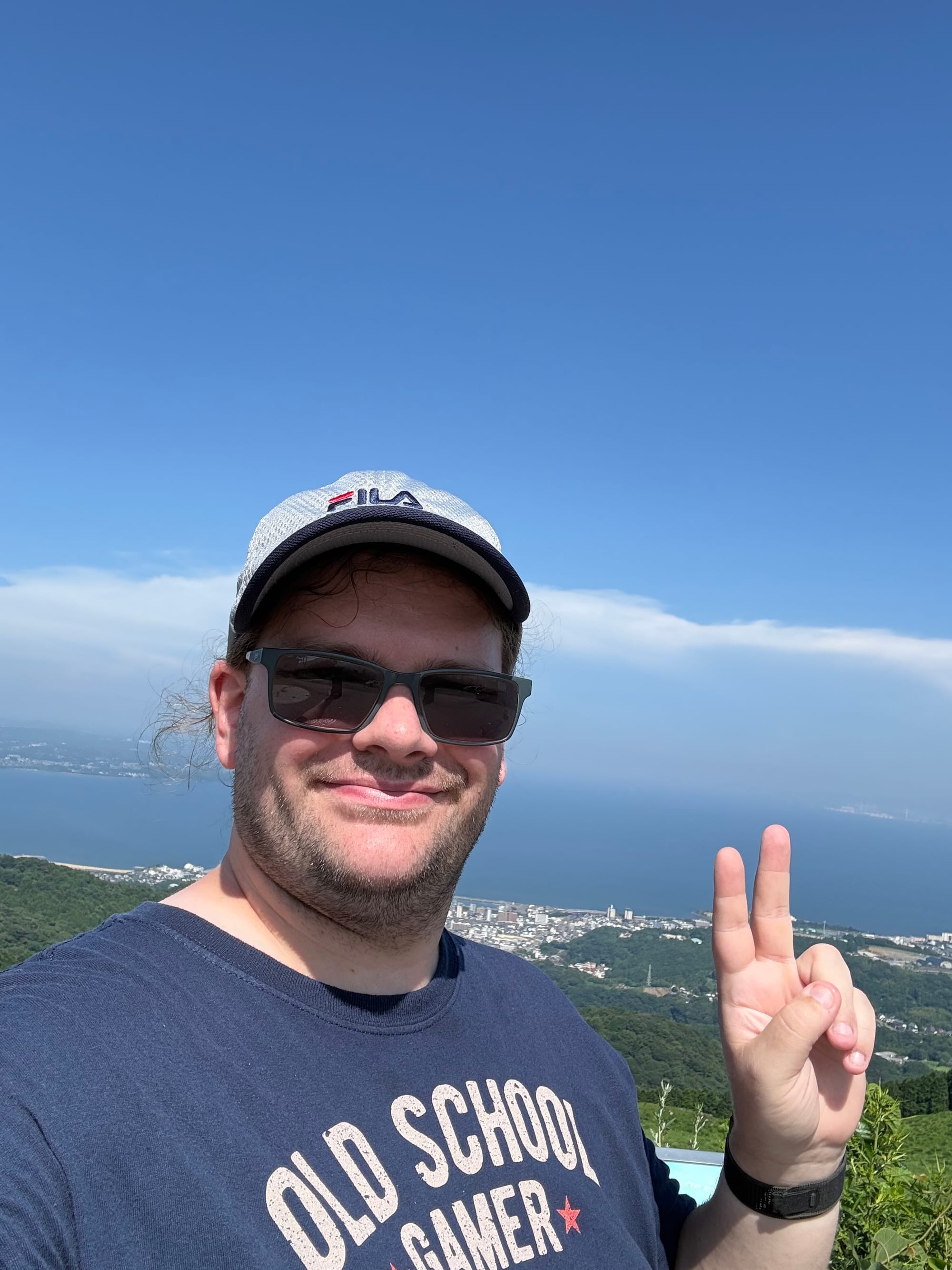 Visitor wearing sunglasses and a cap makes a peace sign at Jūmonjibaru Observatory, with Beppu Bay, the coastline and forested hills stretching into the distance.