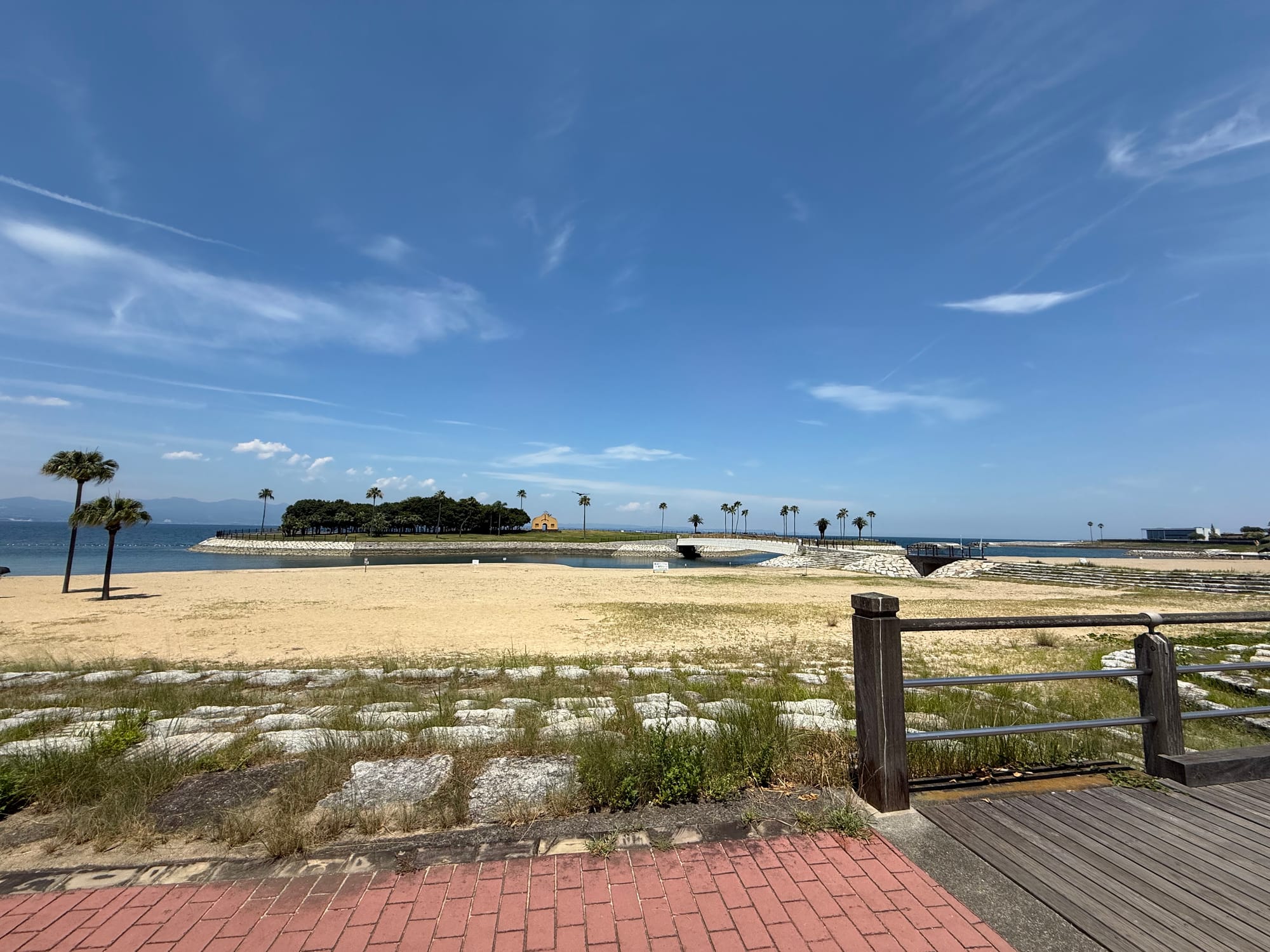 View from Tamoura Beach in Beppu towards Tamoura Isle, with sandy shores, palm trees, a lagoon and calm waters beneath a bright blue summer sky.