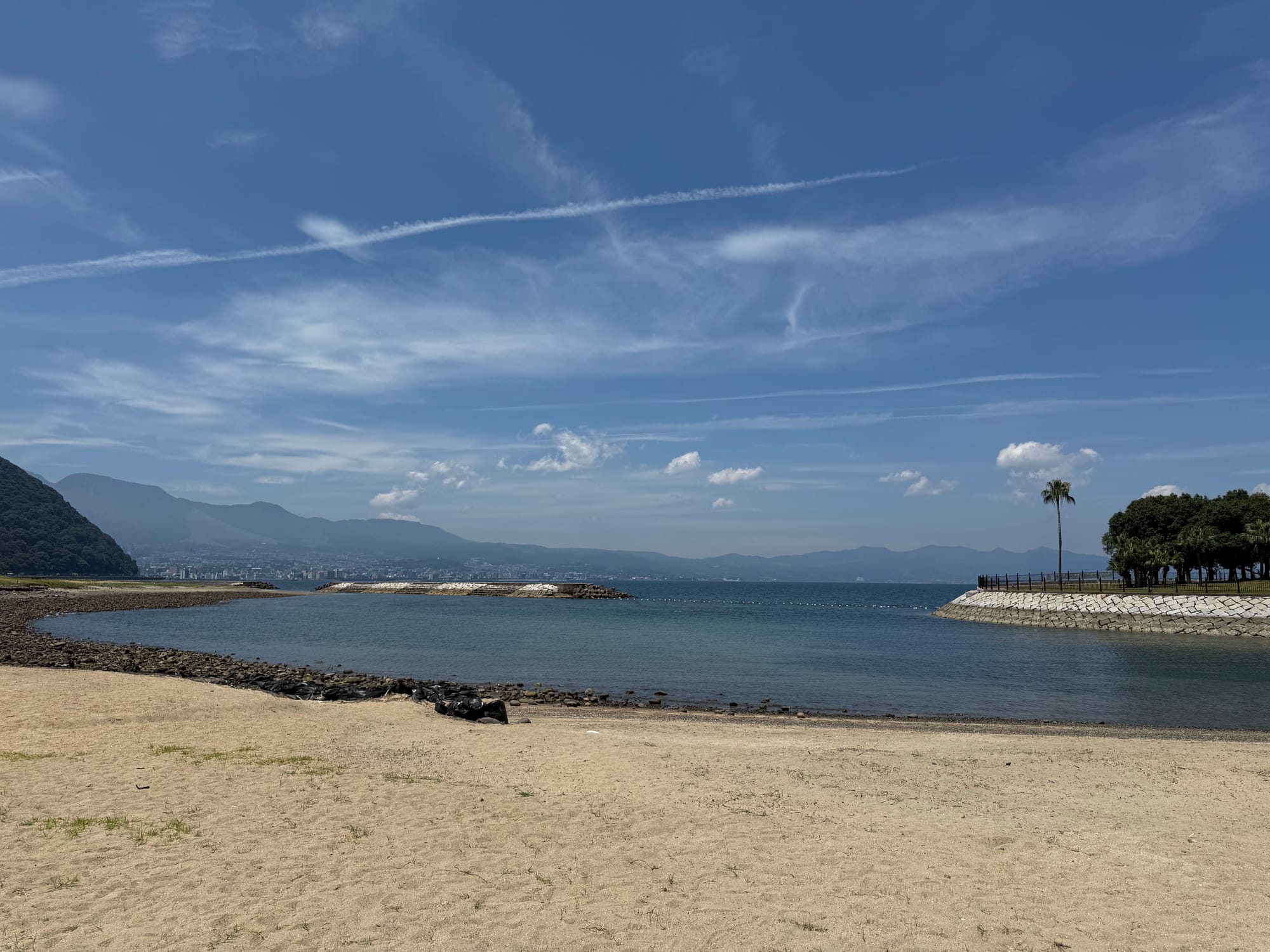 View from Tamoura Beach in Beppu across calm waters and a sandy shoreline, with distant mountains, a breakwater and wispy clouds beneath a bright blue sky.