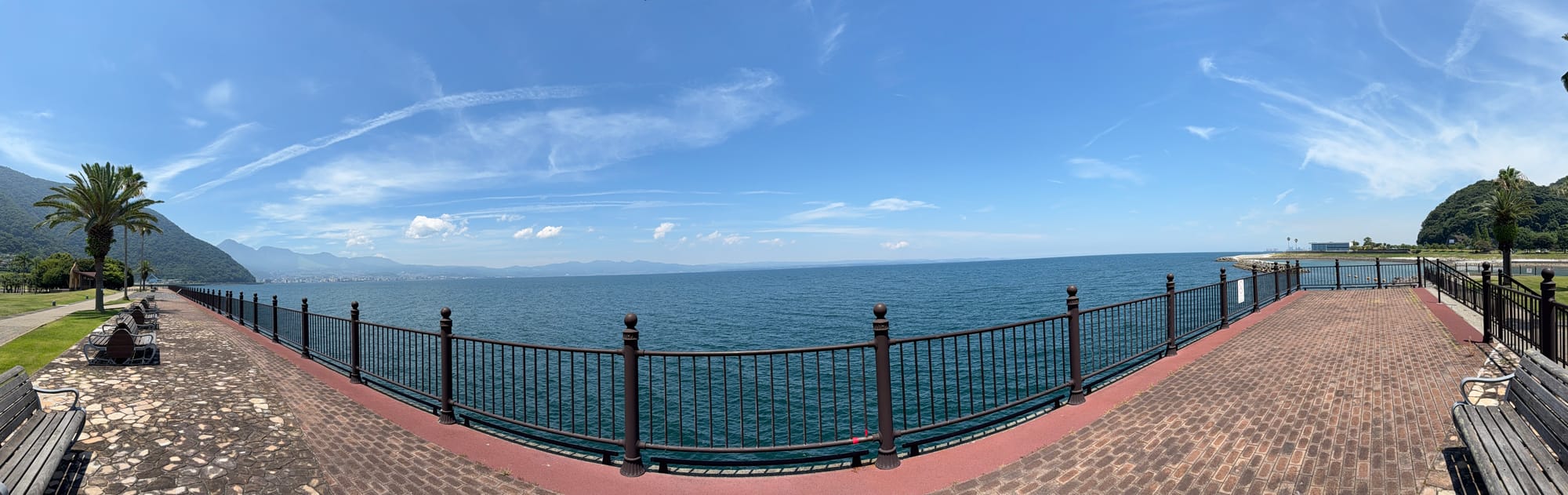 View from Tamoura Isle in Beppu across Beppu Bay, with a waterfront promenade, palm trees, benches, calm blue water and distant mountains beneath a clear sky.