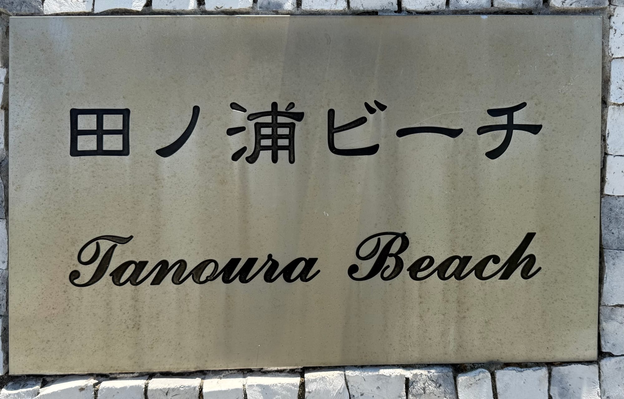 Stone sign for Tamoura Beach in Beppu, displaying the beach name in Japanese and English on a polished plaque set into a white stone wall.