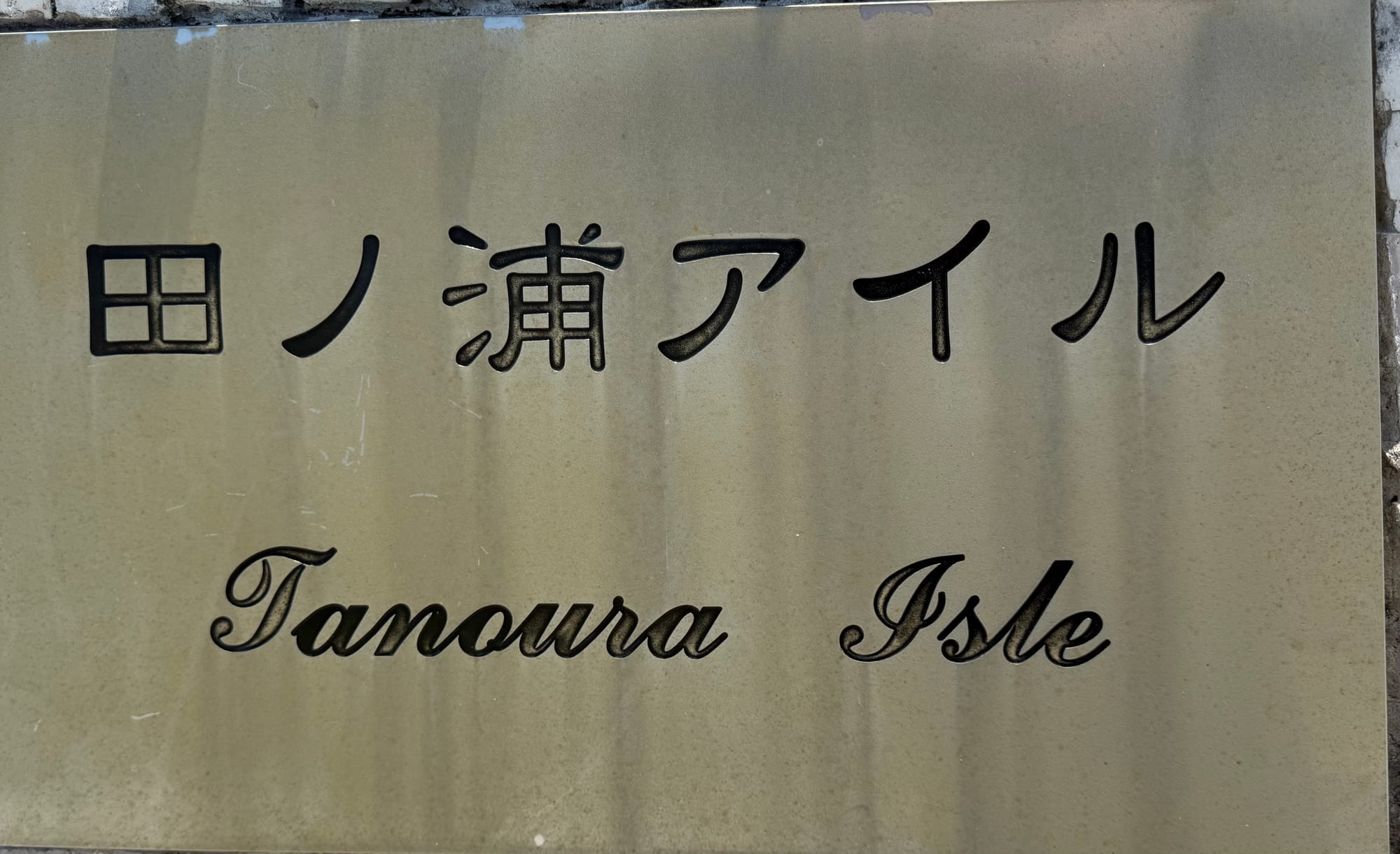 Stone sign for Tamoura Isle in Beppu, displaying the name in Japanese and English on a polished plaque set into a white stone wall.