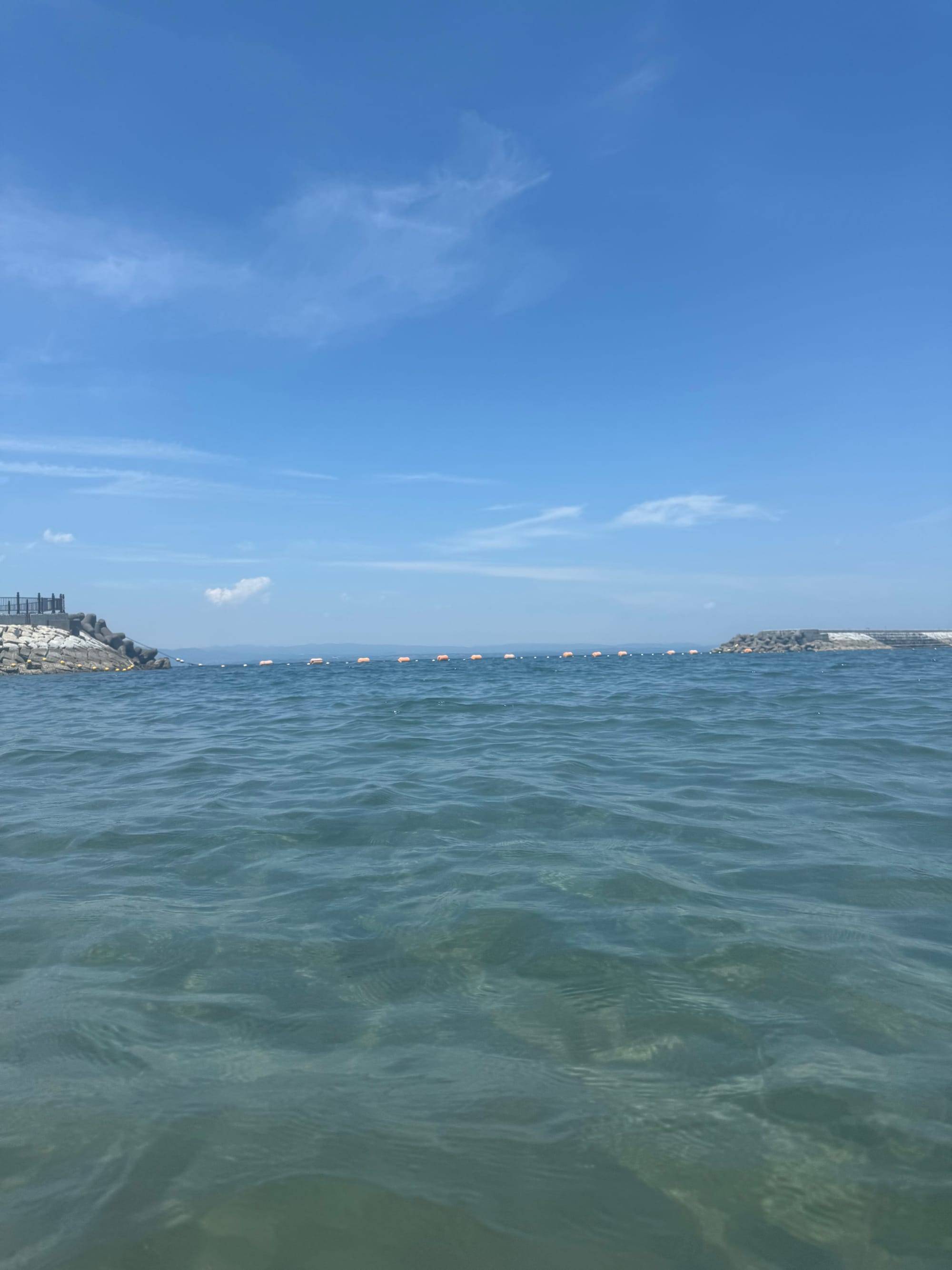 View from the sea at Tamoura Beach in Beppu, with clear water, floating safety buoys, rocky breakwaters and a bright blue summer sky stretching overhead.