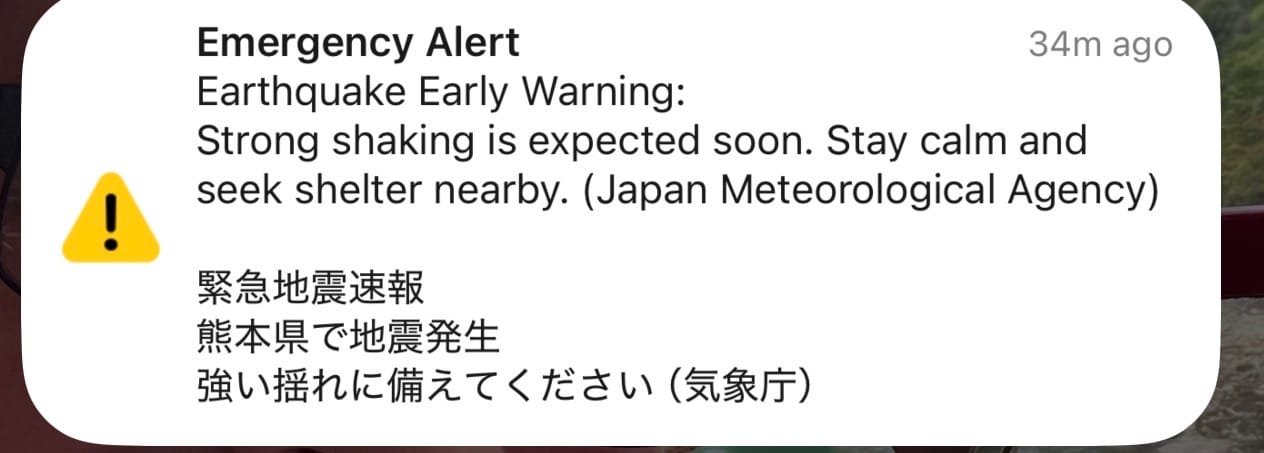 Emergency earthquake warning notification on a smartphone screen in Japan, advising that strong shaking is expected and instructing people to stay calm and seek shelter.