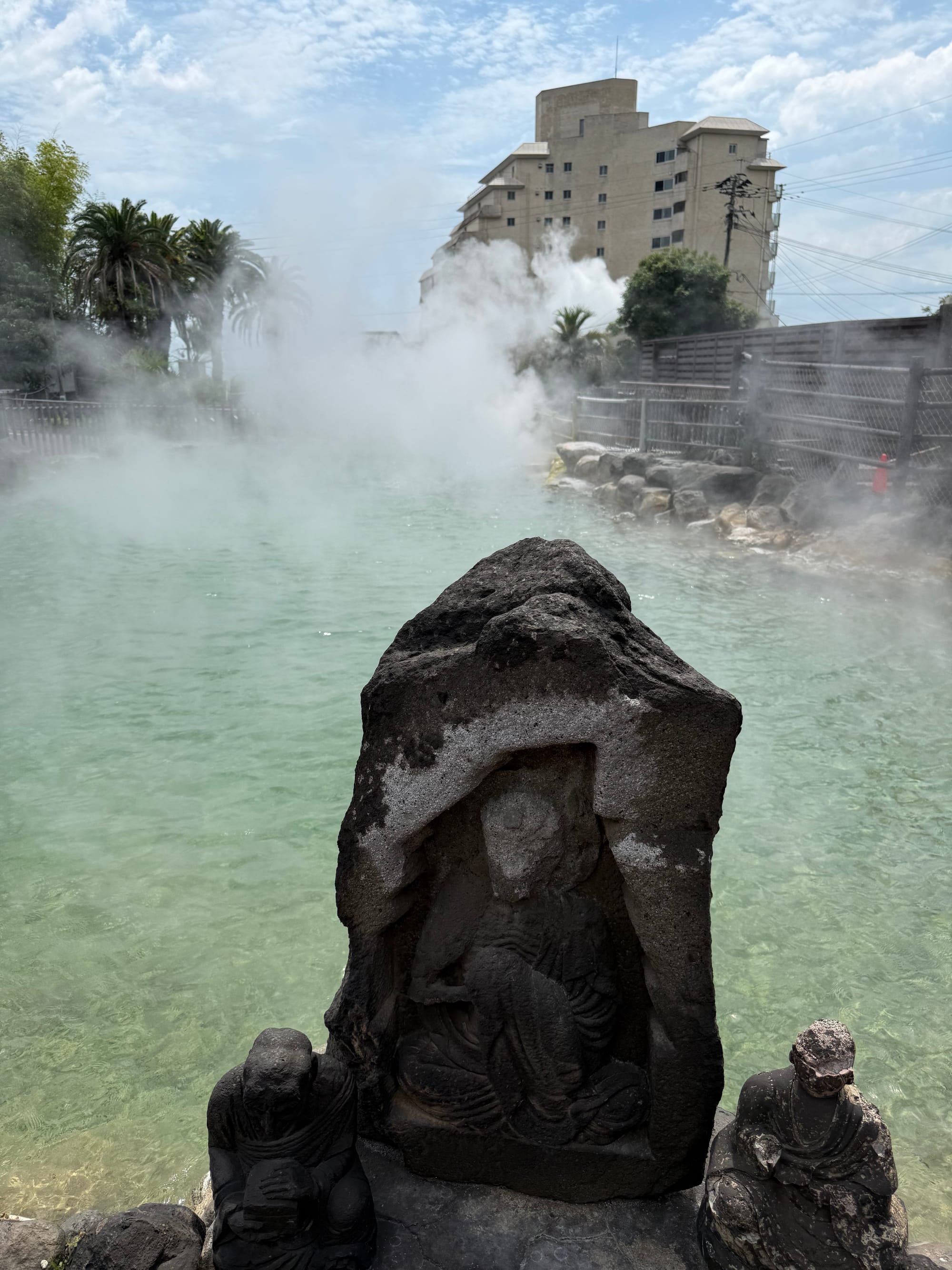 Stone Buddhist statues beside a steaming geothermal pool at Oniyama Jigoku in Beppu, with clear mineral-rich water, rising steam and nearby buildings in the background.
