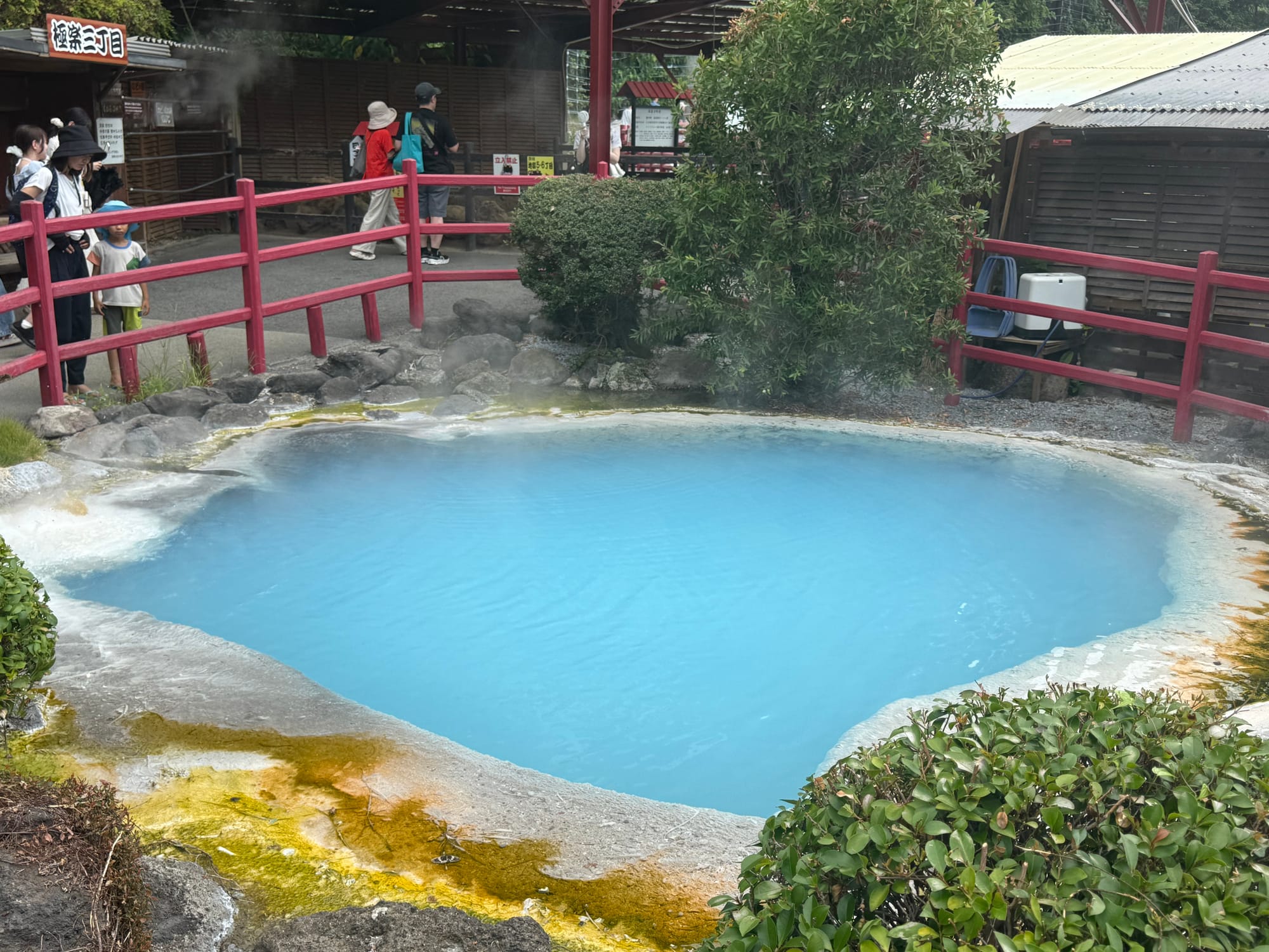 Vivid blue geothermal pond at Kamado Jigoku in Beppu, surrounded by mineral deposits, steam, red railings and landscaped gardens within the famous hot spring attraction.